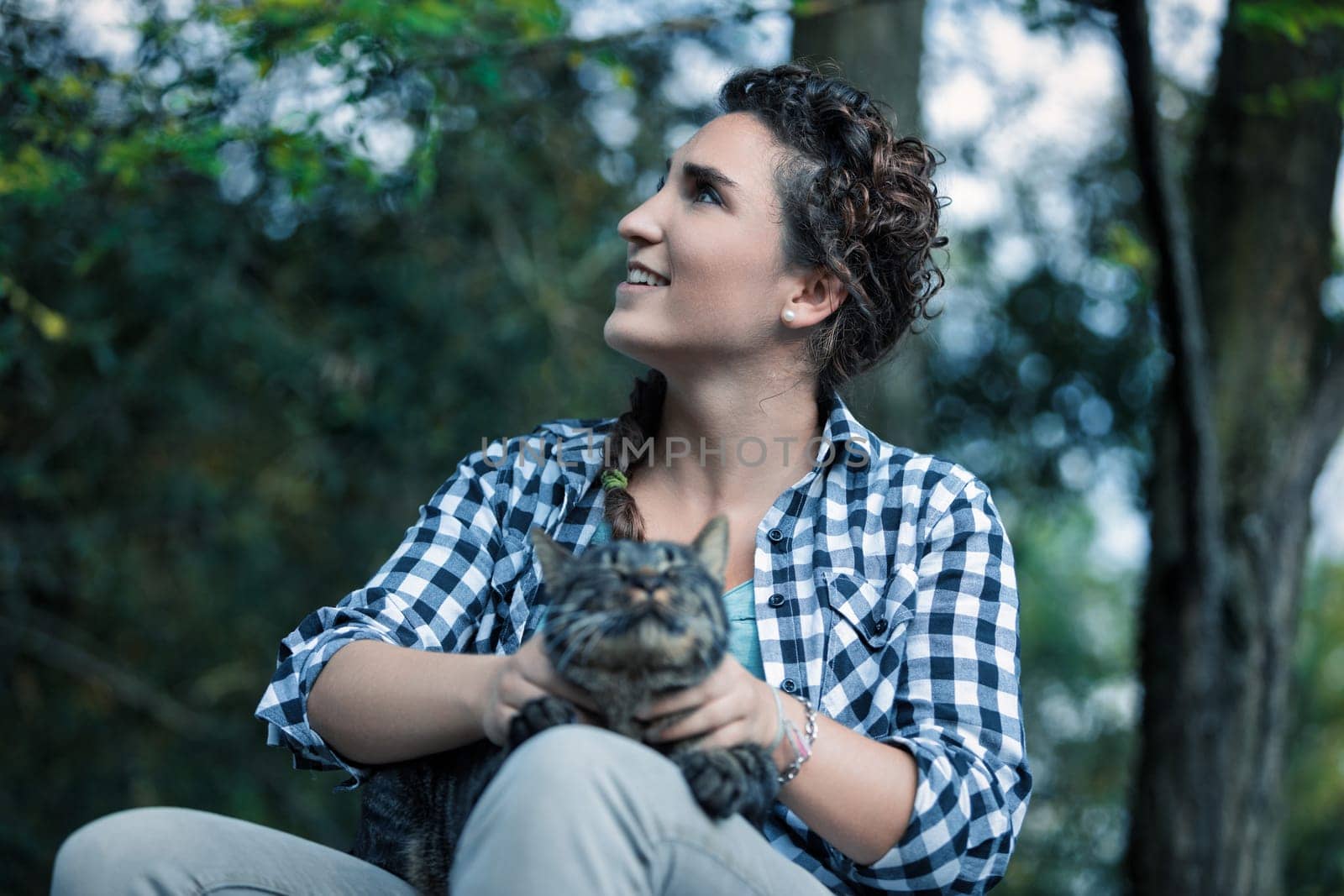 Young woman sits in a forest, holding her tabby cat, looking up with a smile, enjoying nature. They share a moment of love and happiness, surrounded by trees