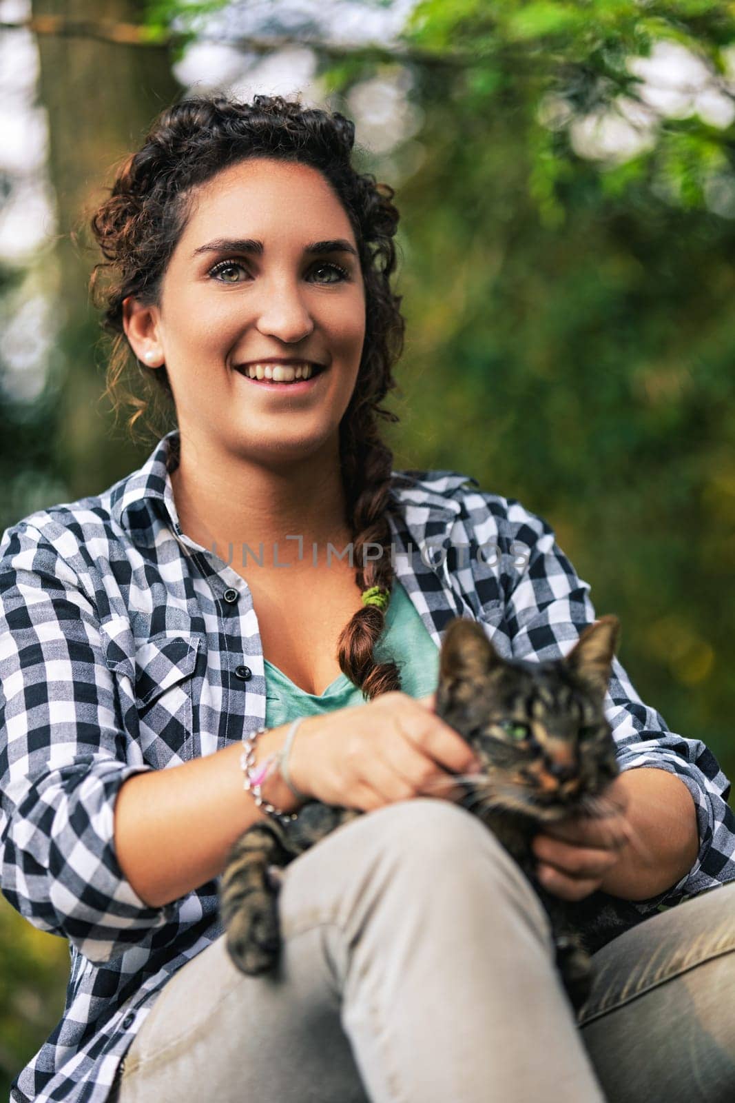 Woman enjoying peaceful moment outdoors with her tabby cat, surrounded by lush greenery. Braided hair, plaid shirt, embodying tranquility. Heartwarming bond between humans and animals