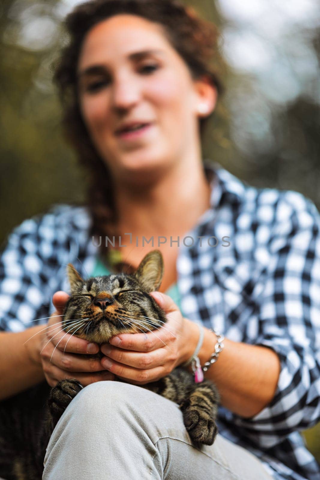 Young woman gently holds a tabby cat's face outdoors, showing love and connection. Smiling, they share a tender moment in nature, reflecting a strong bond