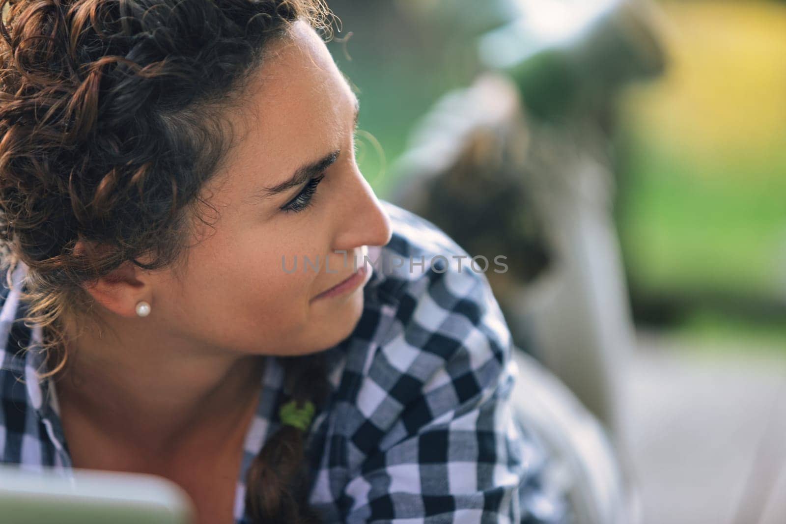 Young woman with curly brown hair, wearing a checkered shirt, lies down and looks away with a pensive expression on her face, in a blurred garden background