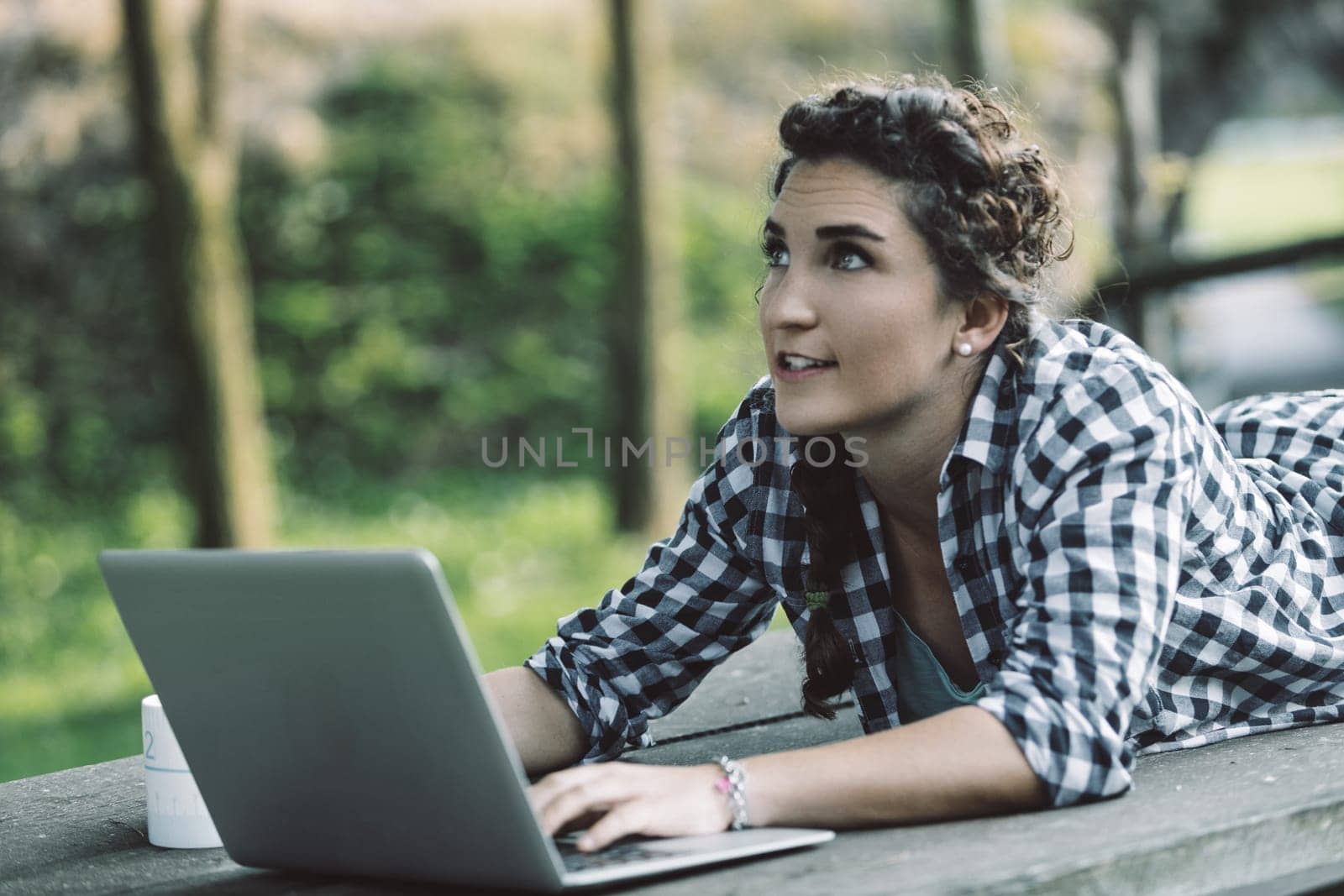 Young woman lying on a table in a park, using a laptop while enjoying the fresh air and natural surroundings, thinking creatively and looking up at the sky