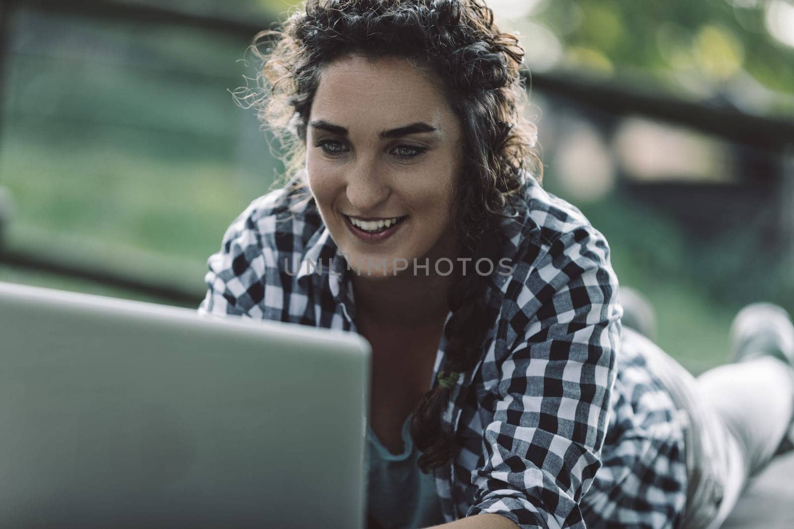 Young woman with curly hair, wearing a checkered shirt, lies on her stomach outdoors, smiling as she works on her laptop, possibly enjoying the fresh air and natural light while staying connected