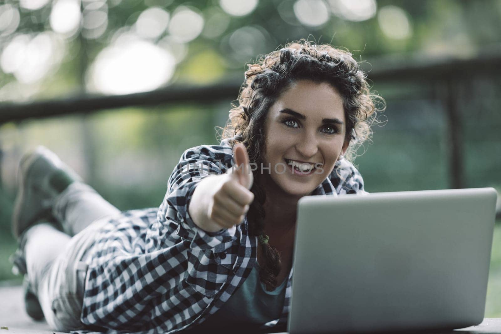 Happy young woman lying in a park, using a laptop while giving a thumbs up, celebrating her success in online education and enjoying the freedom of learning new skills outdoors