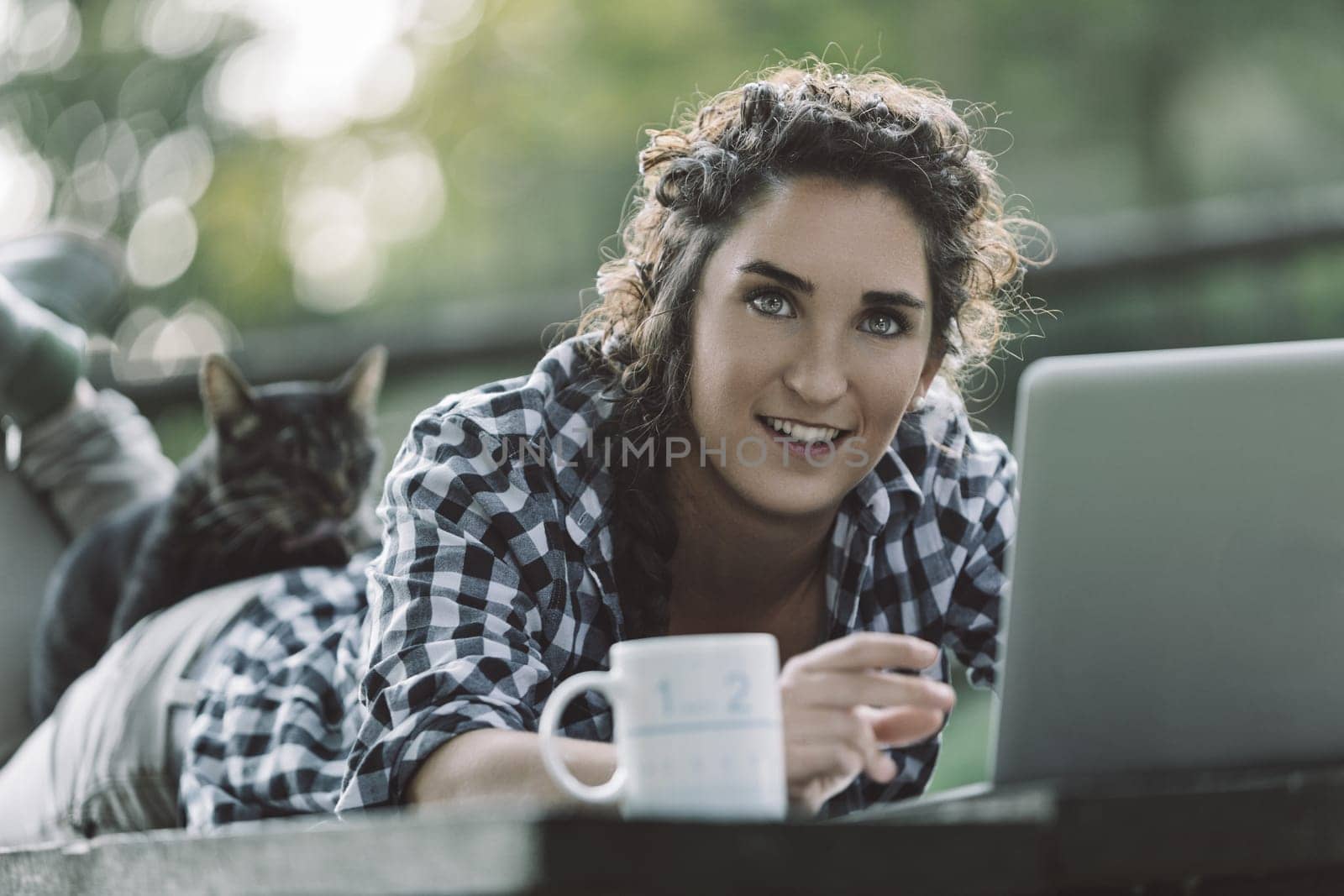 Young woman is lying on her stomach on a wooden table outdoors, working on her laptop with her cat lying on her back and a mug beside her