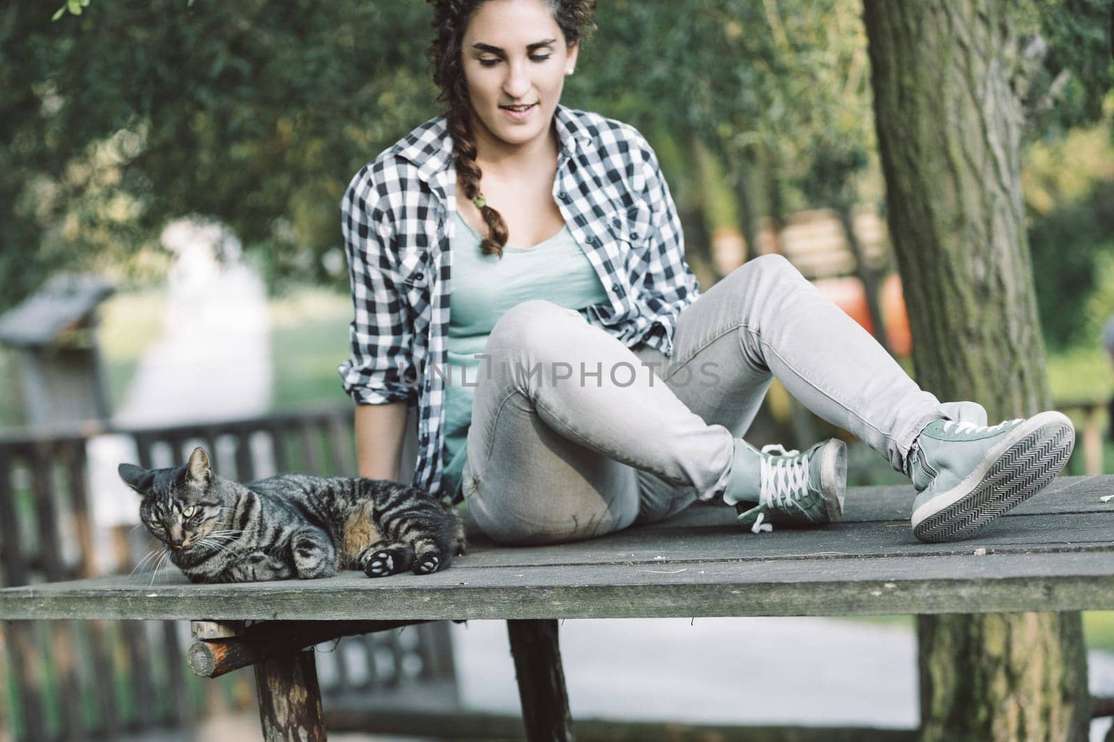 Young woman with braided hair, wearing a plaid shirt and jeans, sits on a wooden table outdoors with her tabby cat resting beside her, enjoying a peaceful moment in nature