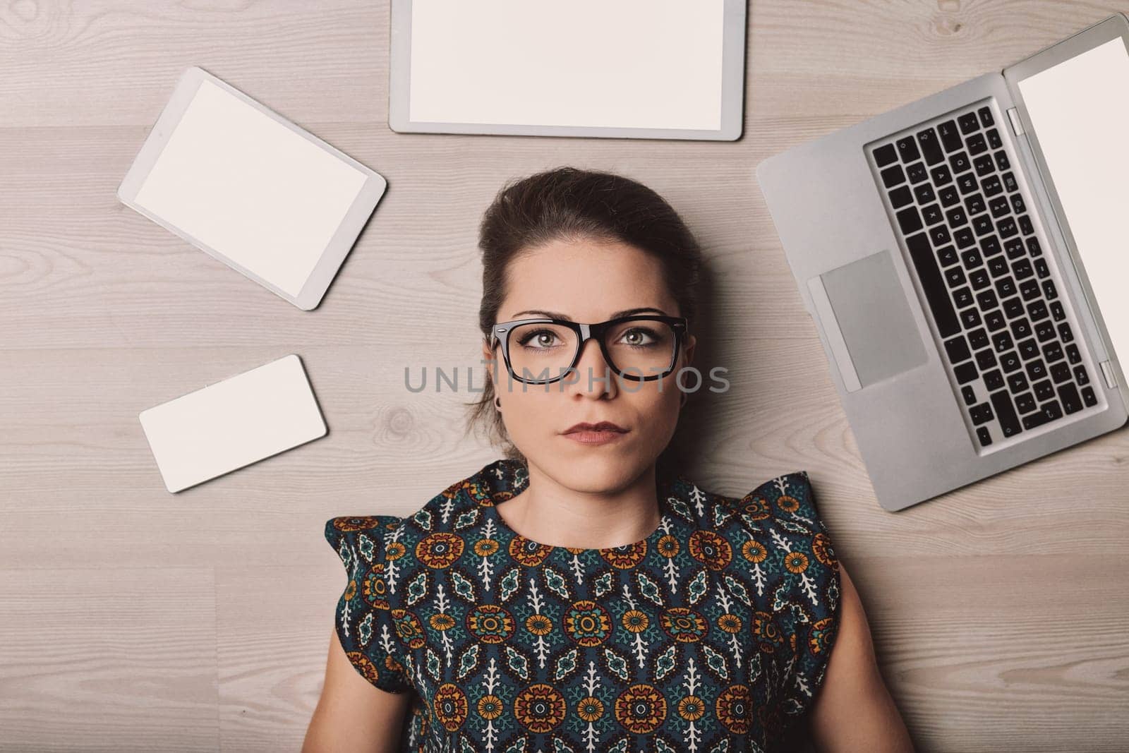 Young woman lying on the floor surrounded by technology devices, looking thoughtful and serious, reflecting in a modern home setting