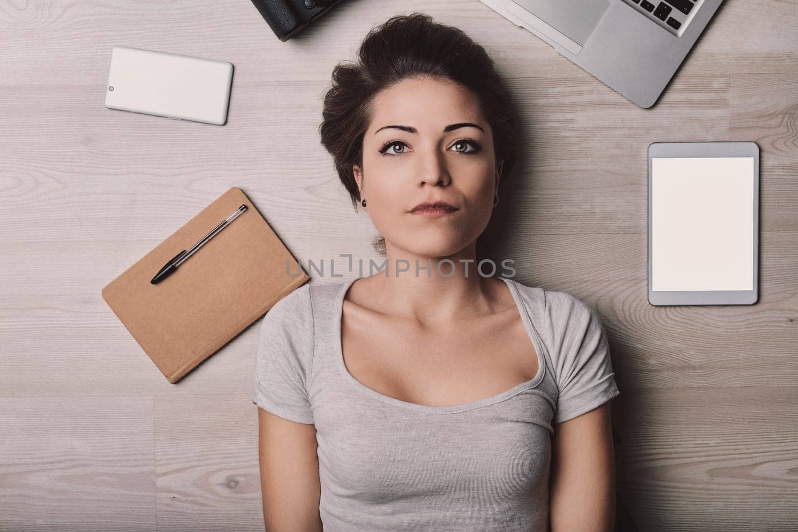 Young woman surrounded by digital devices on a wooden floor, reflecting on modern technology's impact