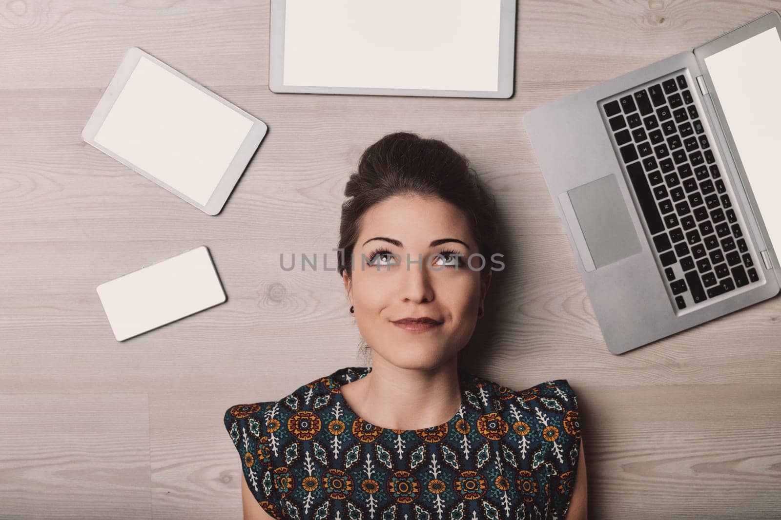 Young woman lies on a wooden floor surrounded by digital devices, lost in thought and gazing up, capturing the essence of the modern digital age