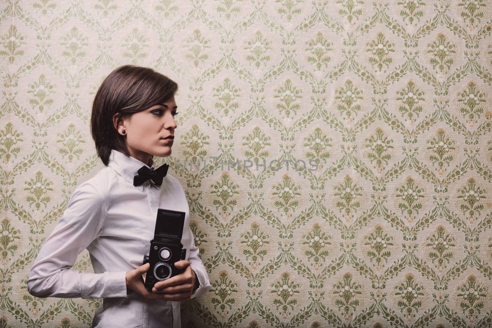 Young female photographer holding a vintage twin-lens reflex camera while posing against a patterned wallpaper, creating a nostalgic and artistic atmosphere