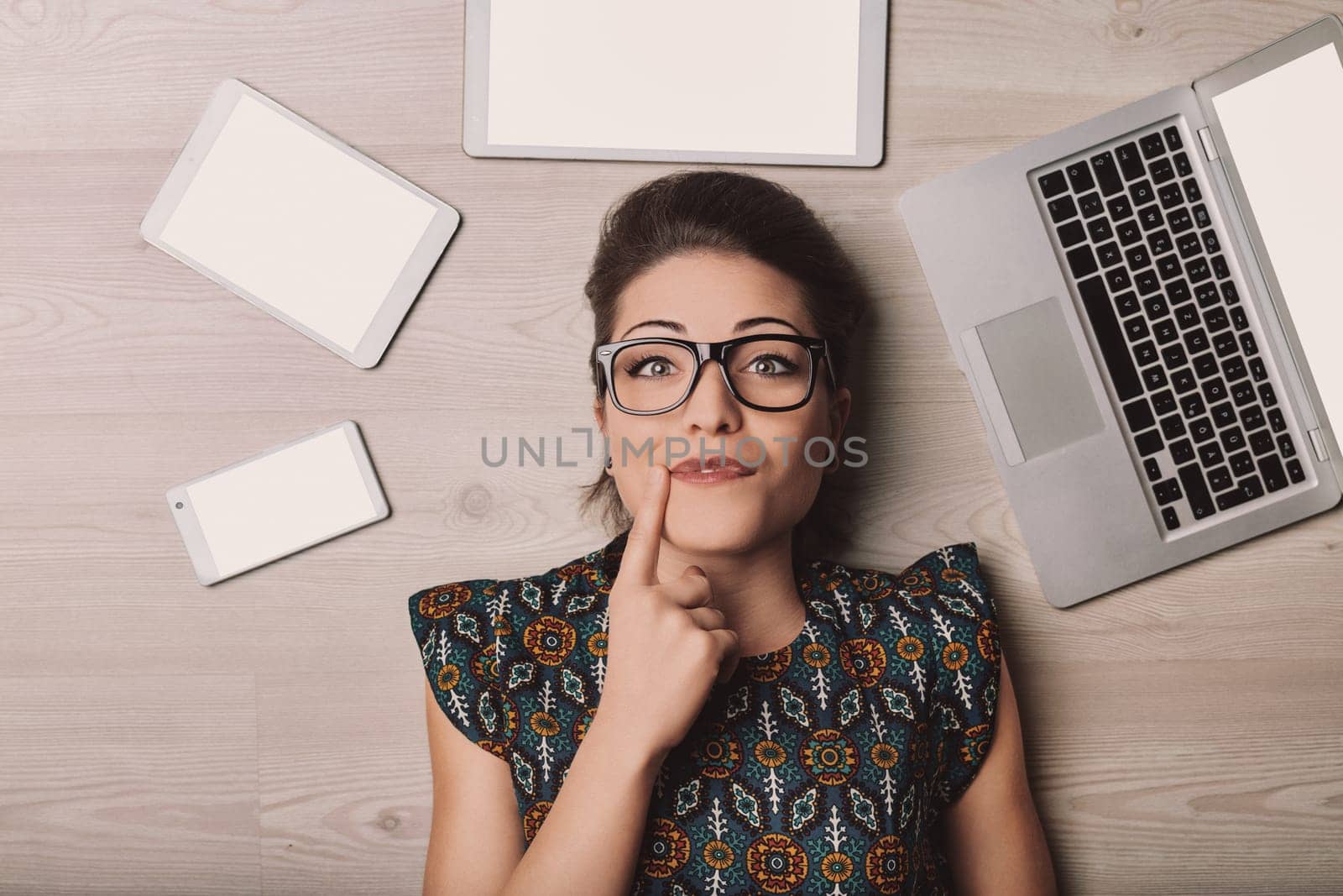 Young woman with eyeglasses lying on the floor surrounded by a laptop, tablet and smartphone, all with blank white screen, touching her chin with her finger and thinking