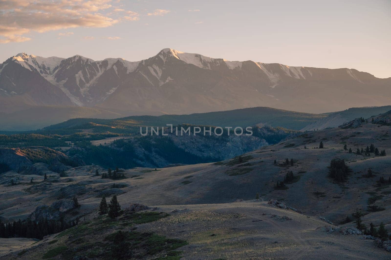 A breathtaking sunset over a rugged mountain landscape, casting warm golden light across hills, trees, and rocky terrain. Altai Mountains