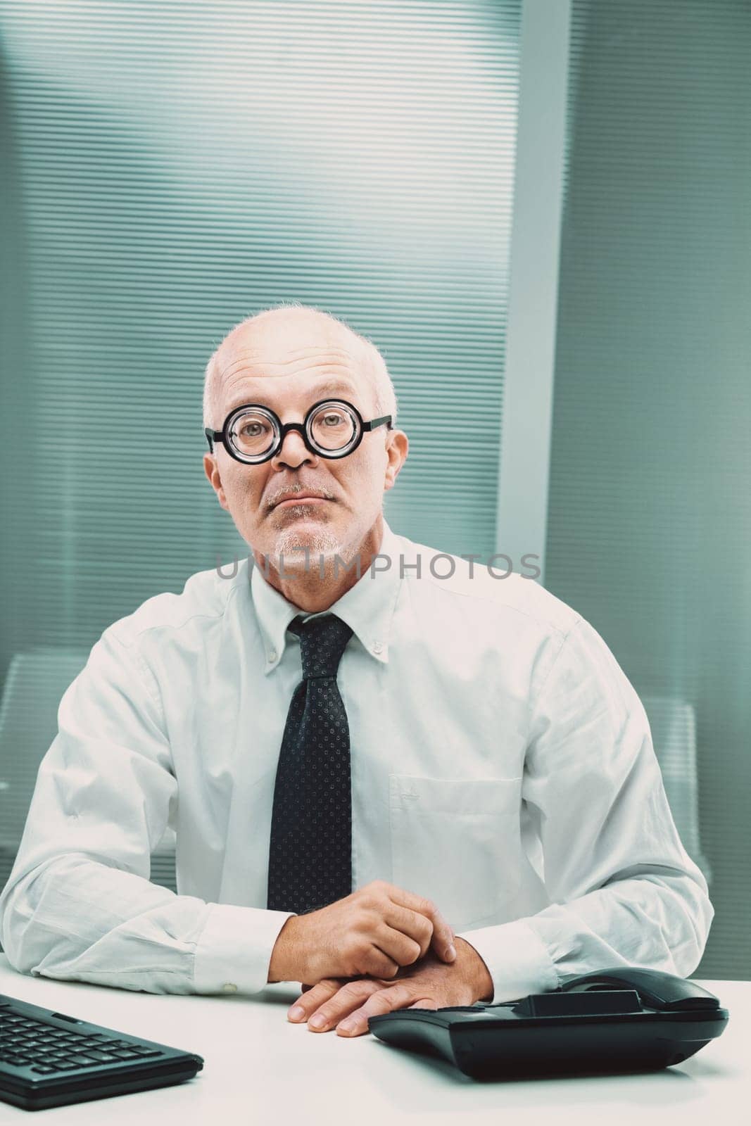 Nerdy businessman with thick glasses sitting at his desk, gazing seriously at the camera while surrounded by office essentials, conveying a professional and focused demeanor