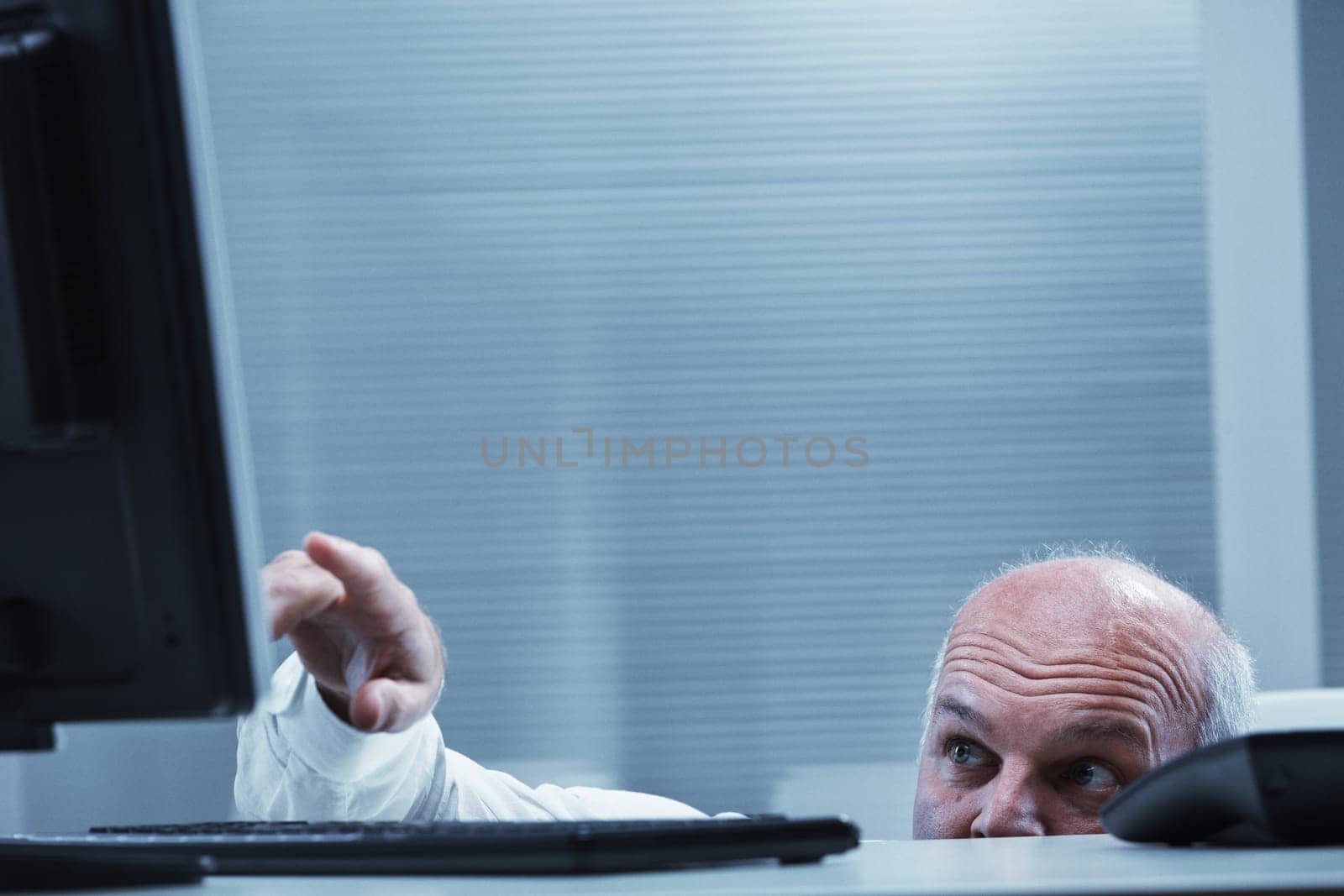 Mature businessman hiding behind his desk, pointing at the computer screen with a curious expression, immersed in the world of technology within a corporate office setting