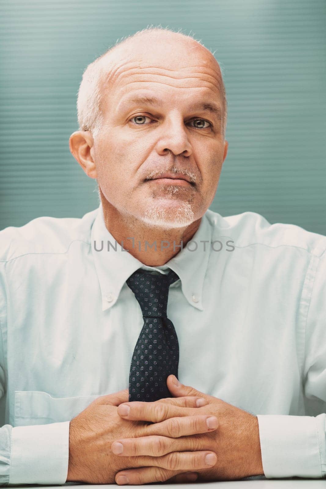 Portrait of a confident mature businessman sitting at his desk, looking directly at the camera with his hands clasped in front of him, wearing a white shirt and tie