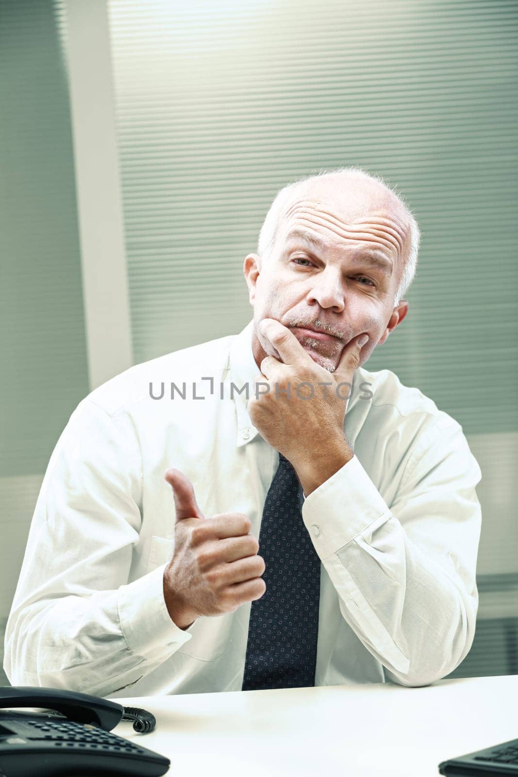 Mature businessman sits at his desk, hand on chin in thought, while giving a thumbs-up gesture, conveying a sense of thoughtful approval or cautious optimism