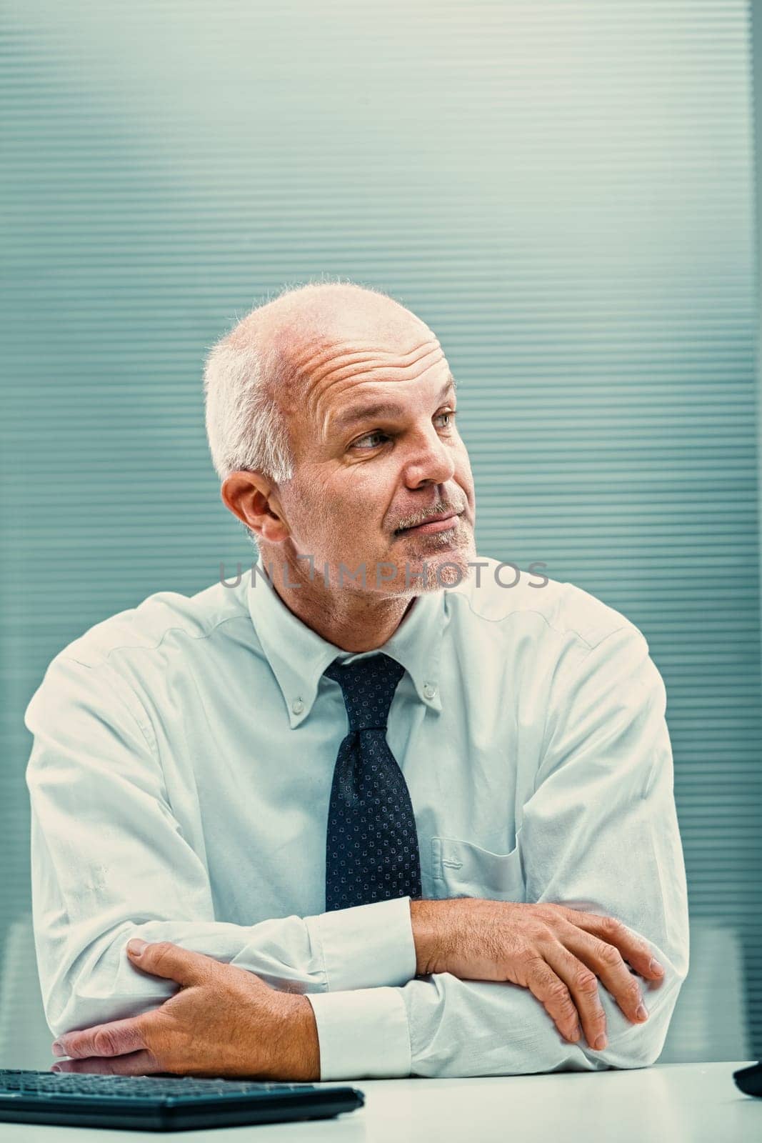 Thoughtful mature businessman sitting at his desk with arms crossed, looking away in contemplation, perhaps considering a new business strategy or reflecting on a challenging decision