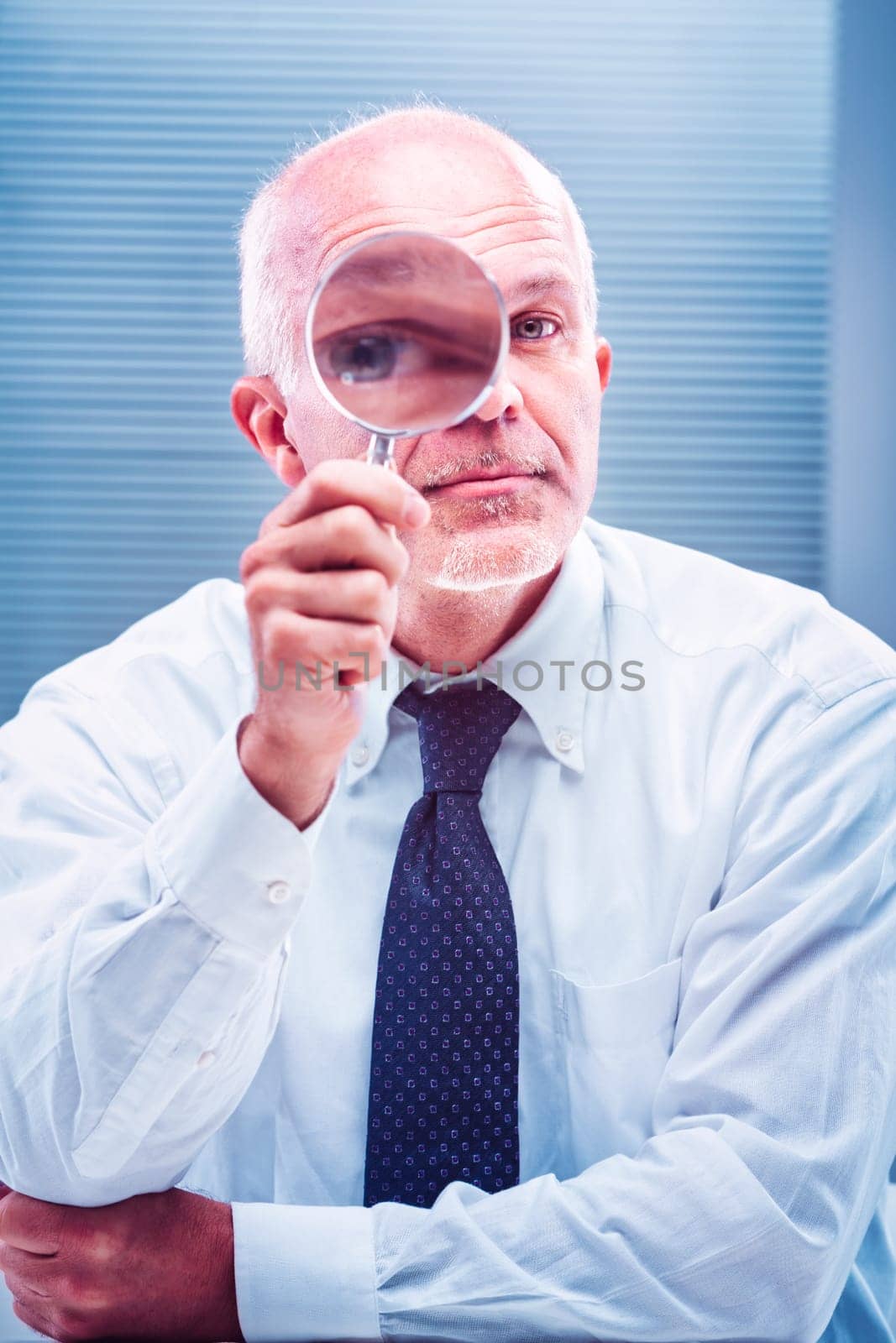 Mature businessman holding a magnifying glass over his eye, looking closely at something, with arms crossed, wearing a light blue dress shirt and tie, in an office setting