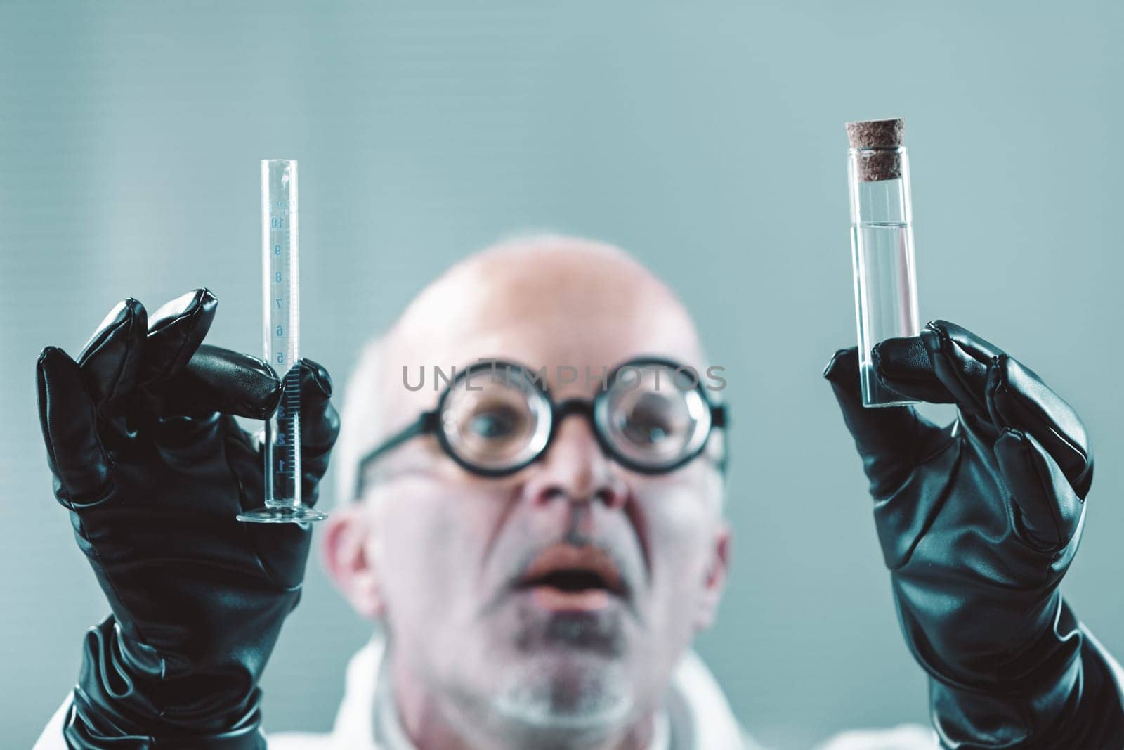 Scientist wearing black gloves holds two test tubes, a graduated cylinder and a corked vial, with a surprised expression, conducting an experiment in a laboratory setting