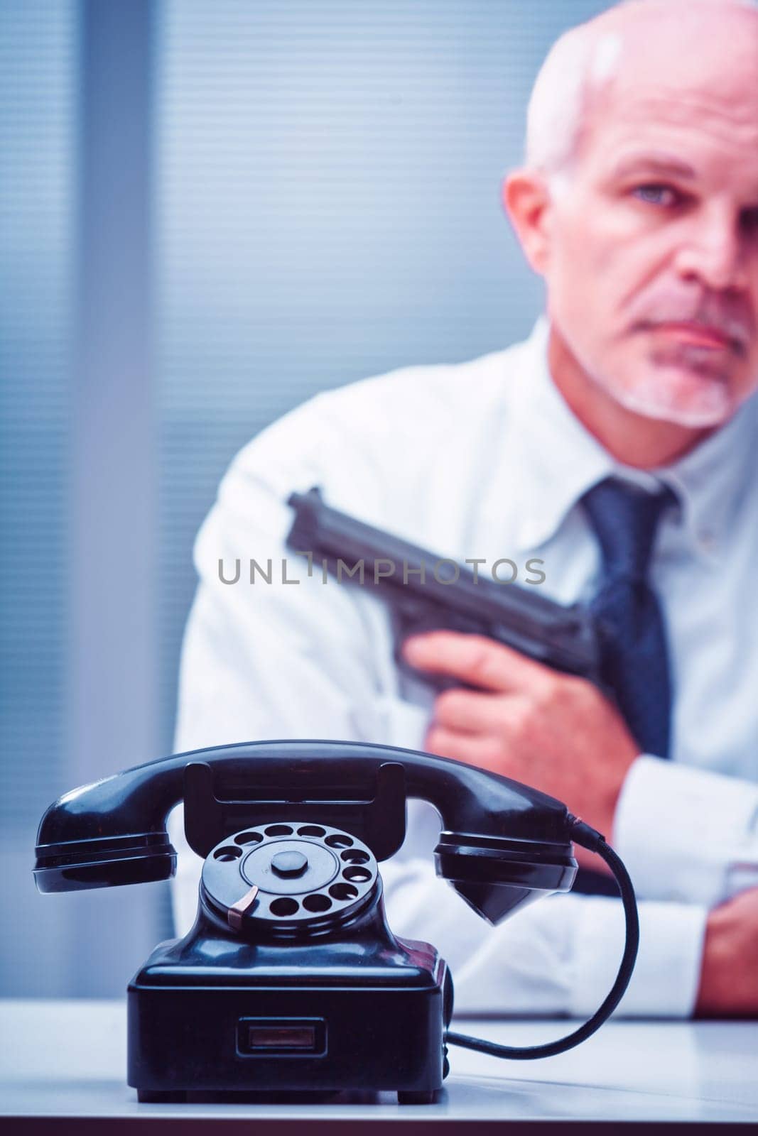 Mature businessman holding a gun behind a vintage telephone on a desk, creating an atmosphere of tension and danger in an office setting, suggesting a serious conflict