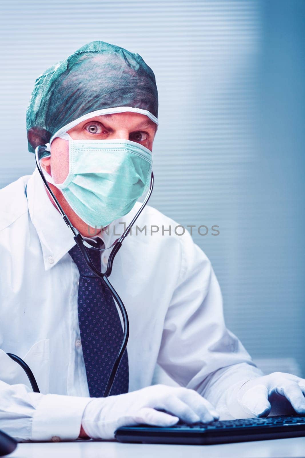 Doctor wearing a surgical mask, hairnet, and gloves, typing on a keyboard while making eye contact with the camera, showcasing a stethoscope around his neck in a clinical setting