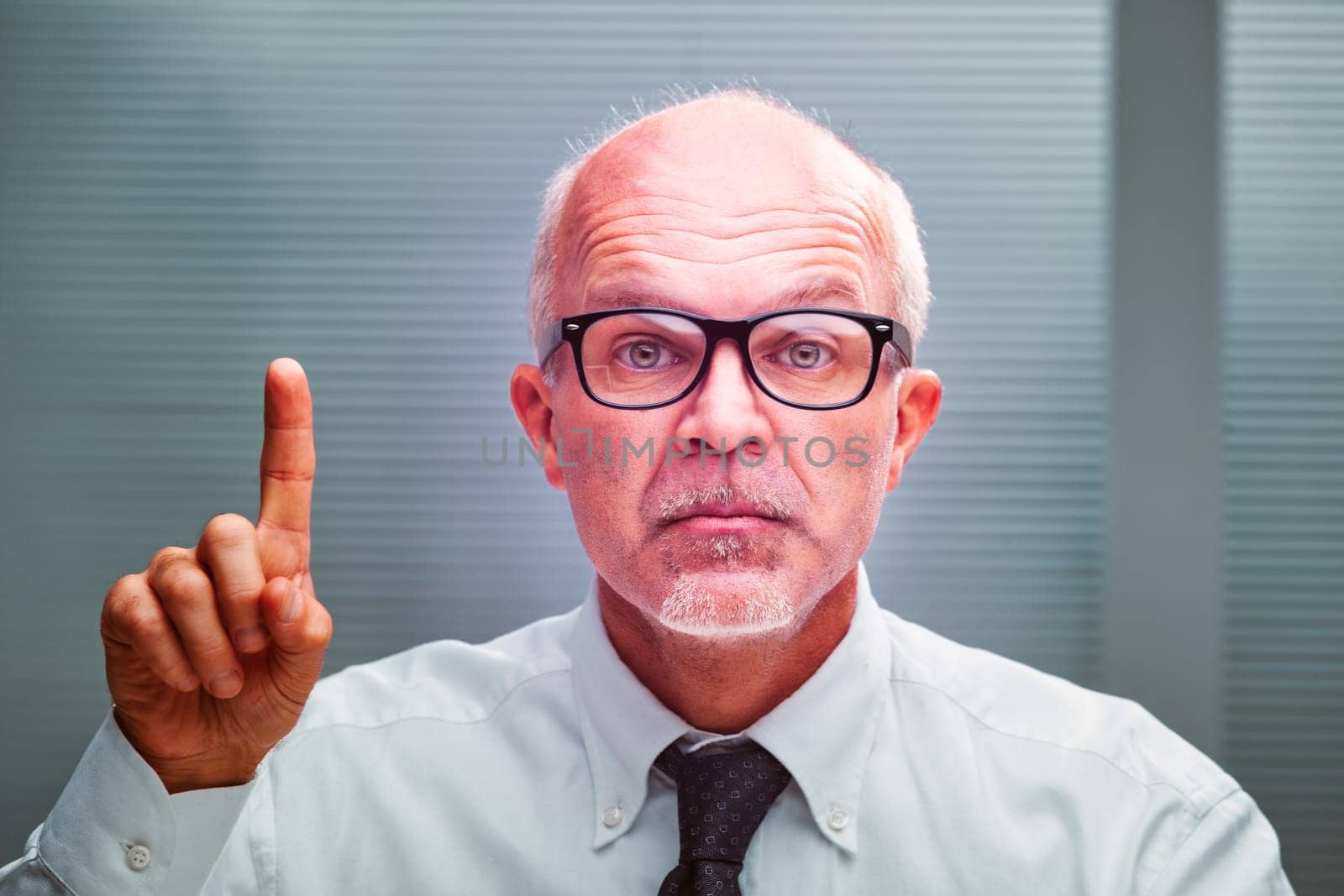 Mature businessman raising his index finger in a warning gesture, exuding seriousness while wearing a crisp white shirt and tie in a professional office setting