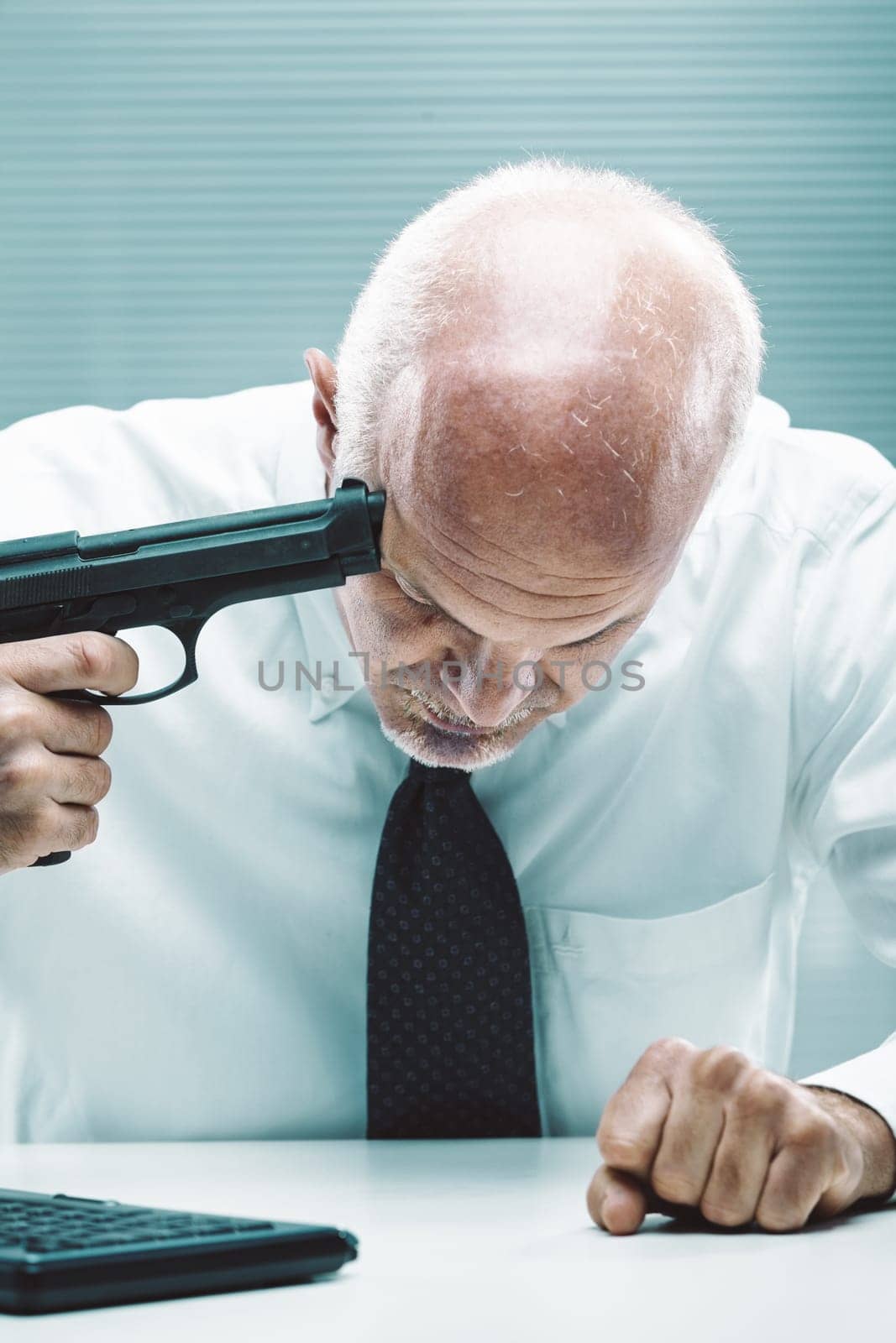 Senior businessman pointing a gun to his head while sitting at his desk in the office, he is stressed and desperate because of overwork and financial problems