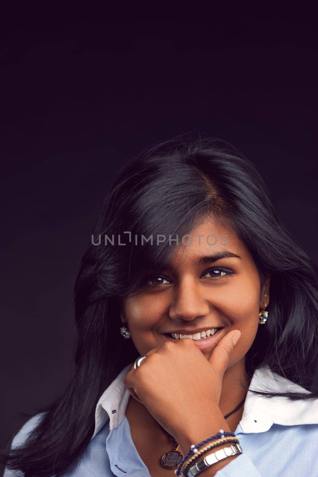 Portrait of a cheerful young indian woman with long black hair, wearing a light blue shirt, smiling and holding her chin with her hand, against a dark background