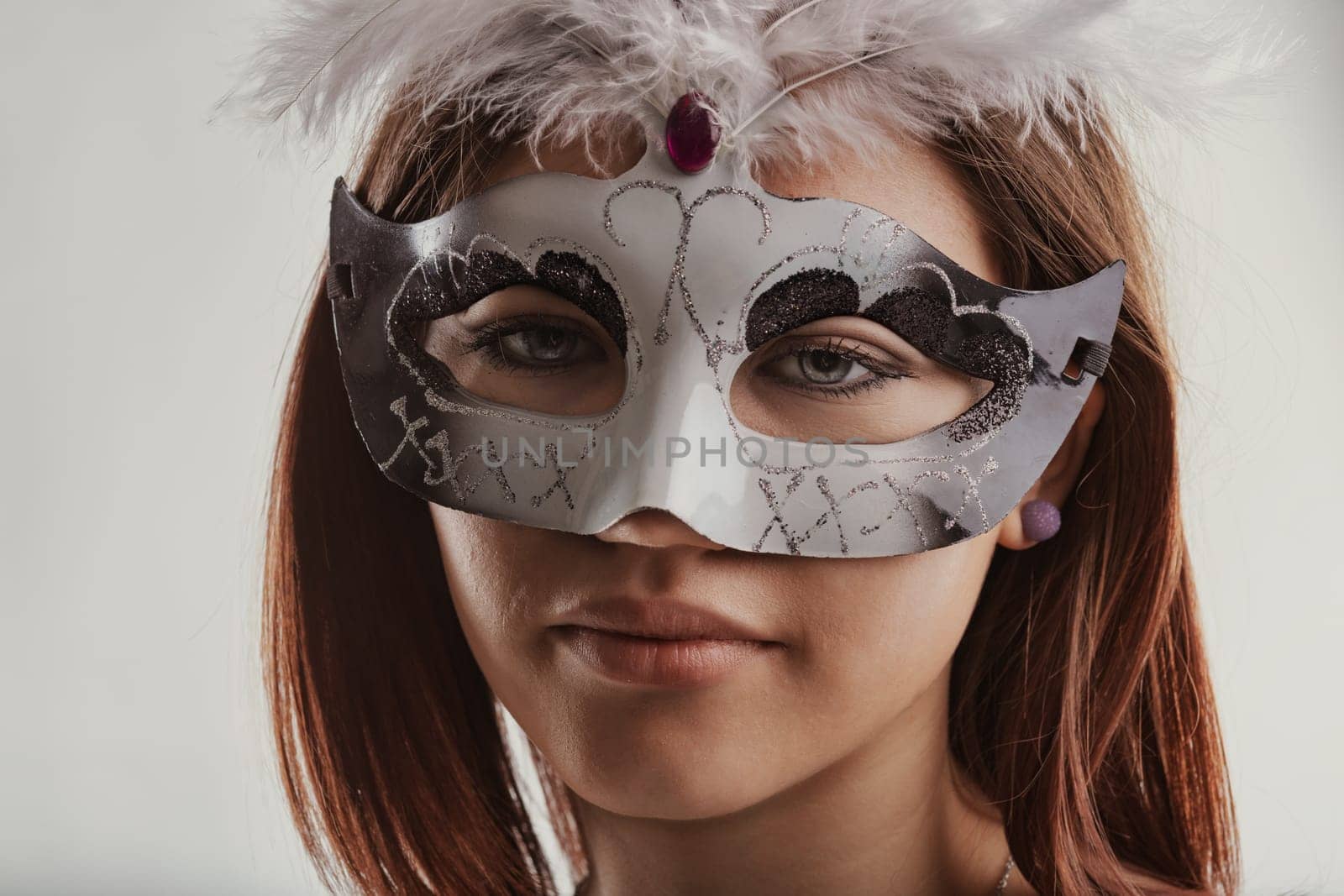Portrait of a young caucasian woman wearing an elegant venetian mask adorned with white feathers, gazing confidently at the camera while posing in a studio against a crisp white background