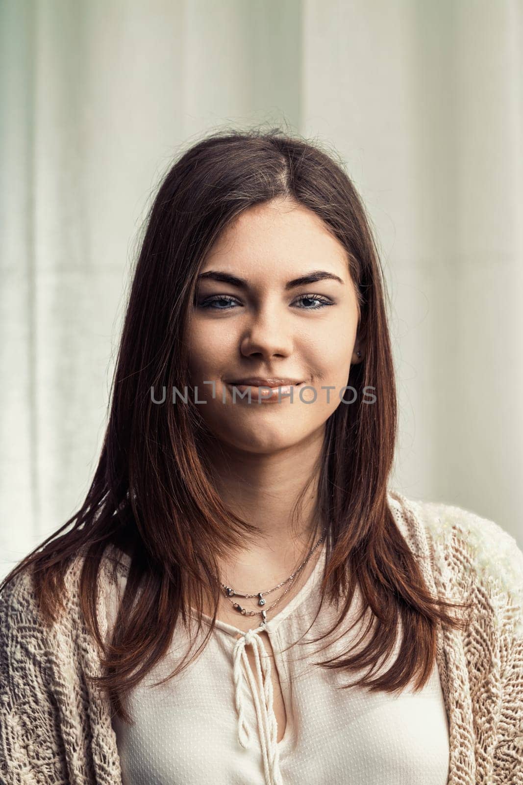 Portrait featuring a beautiful young woman with long brown hair and captivating blue eyes, smiling confidently at the camera, radiating happiness and positivity in a casual white shirt and cardigan