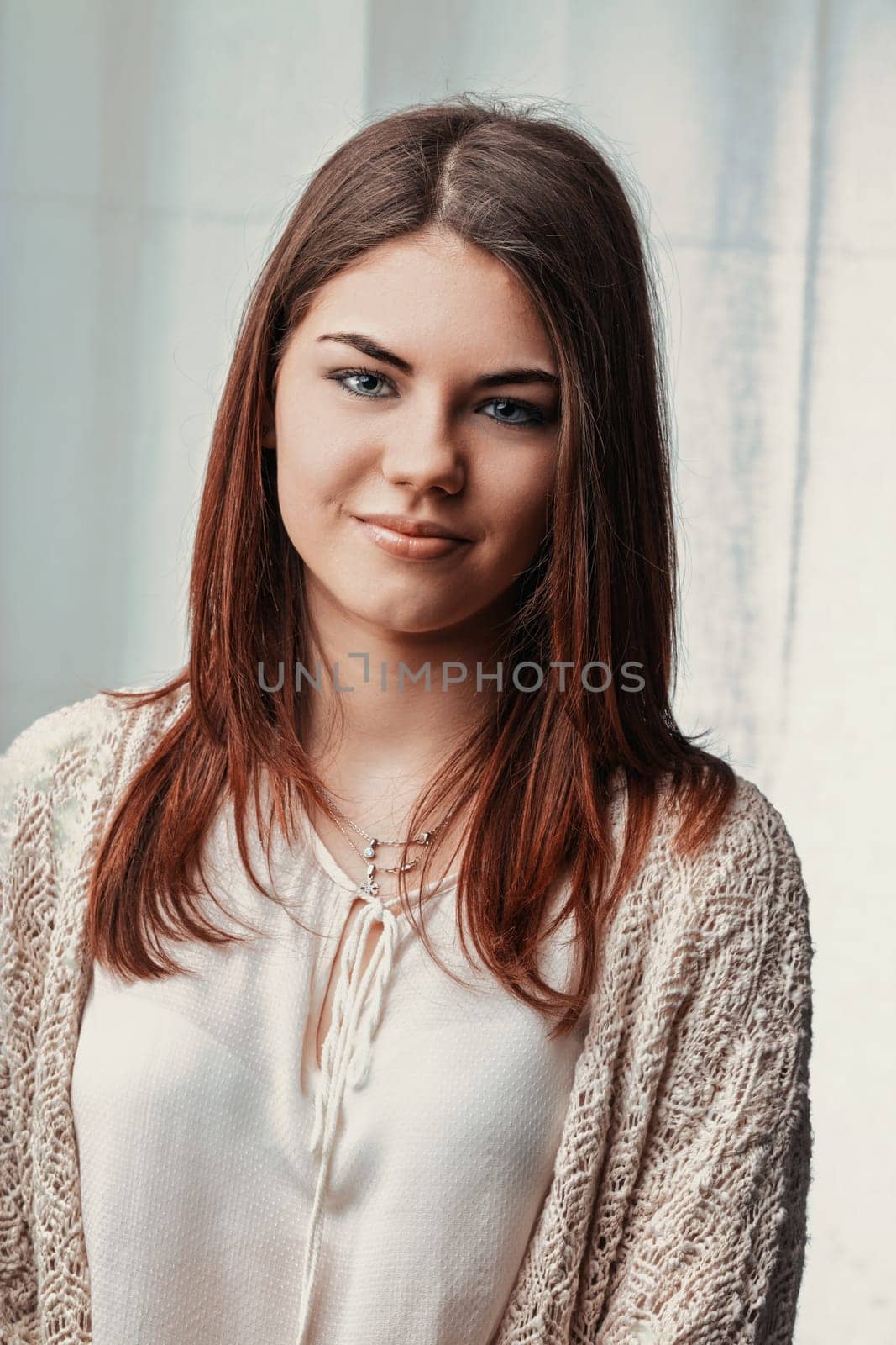 Portrait of a beautiful young woman with auburn hair and blue eyes smiling confidently at the camera, wearing a light beige cardigan and white blouse