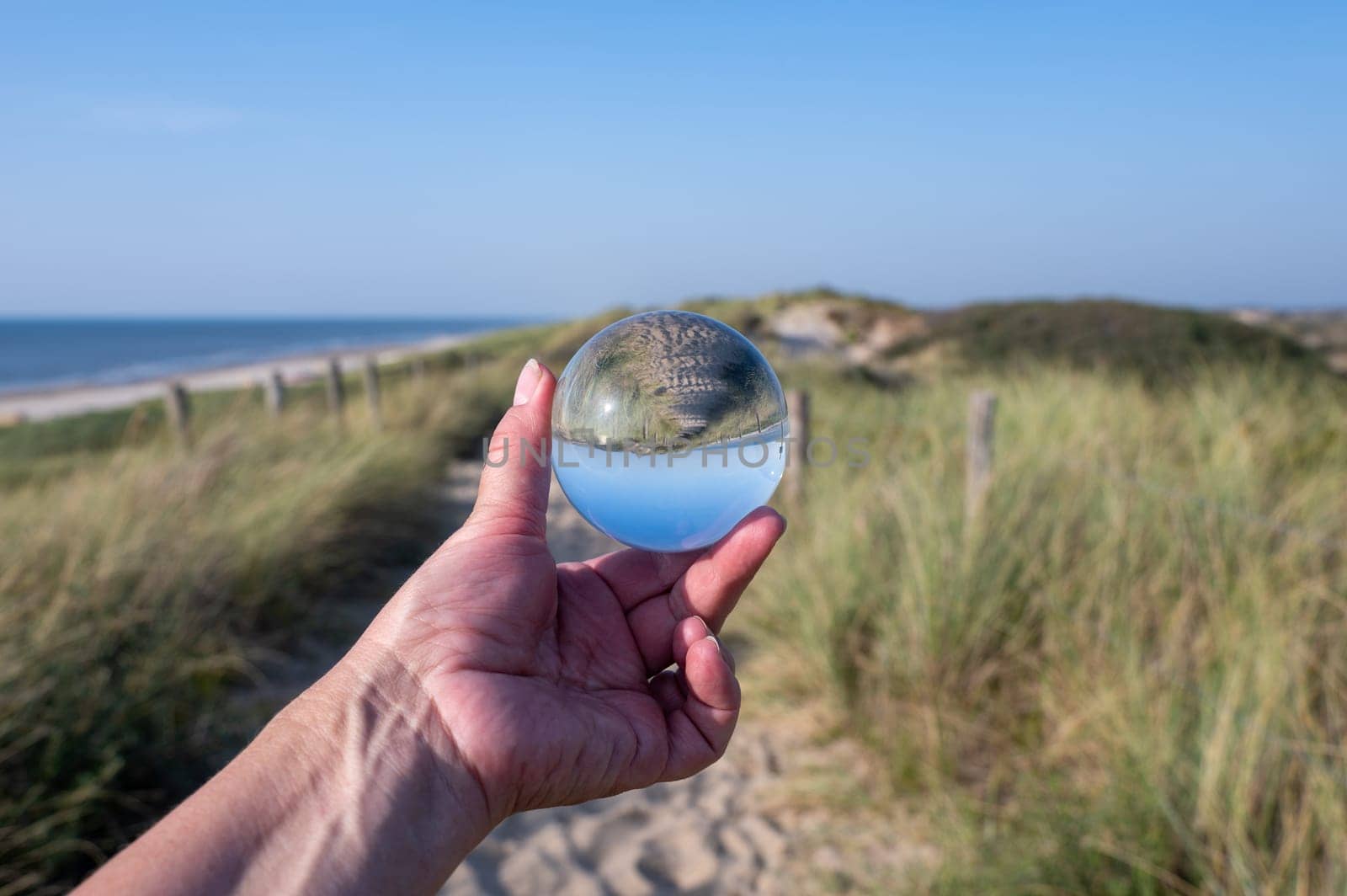 A human hand holds a glass ball, in the ball a sand dune, a footpath, blue sky and the sea are reflected