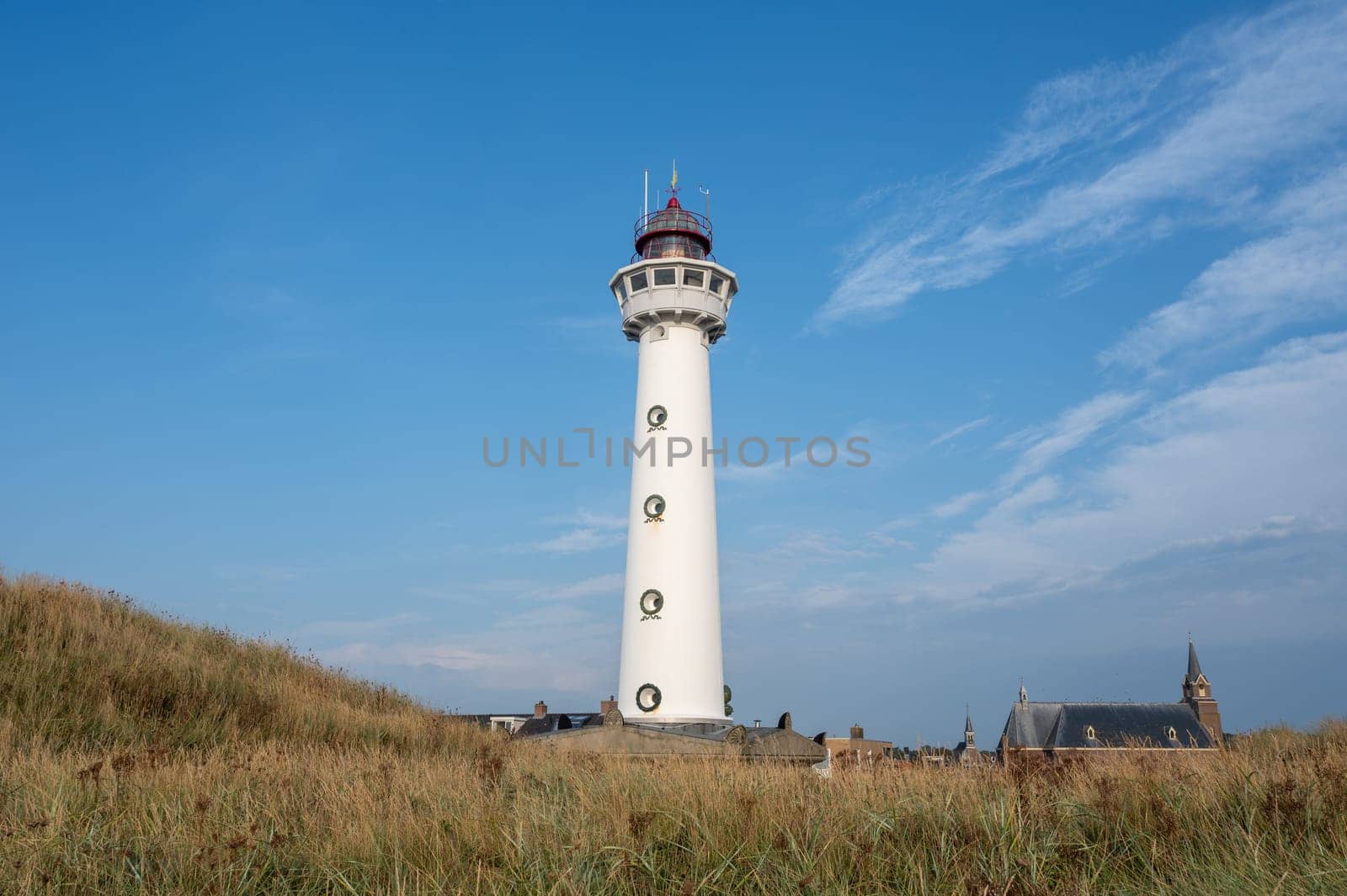 The approx. 28 m high white lighthouse J.C.J. van Speijk in Egmond aan Zee, North Holland, Netherlands with the Catholic St. Agnes Church, beach grass and sand dunes