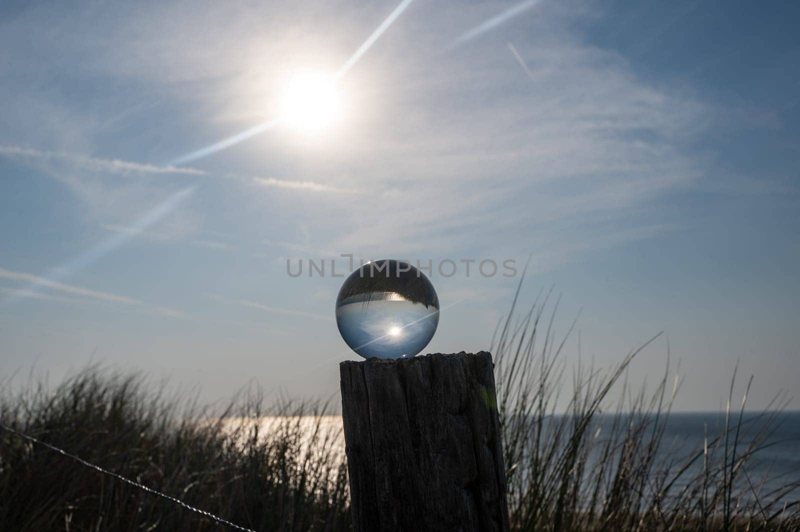 Glass ball lies on a wooden post, beach grass in the background, the sea, the sun and the blue sky are reflected in the ball
