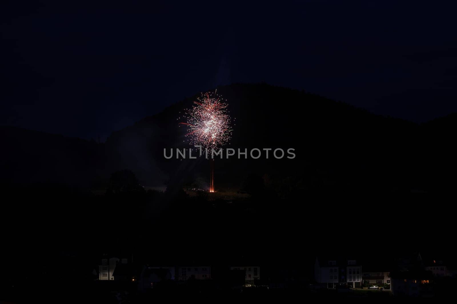 A firework rocket in the sky on a mountain at night, a few illuminated houses in the foreground