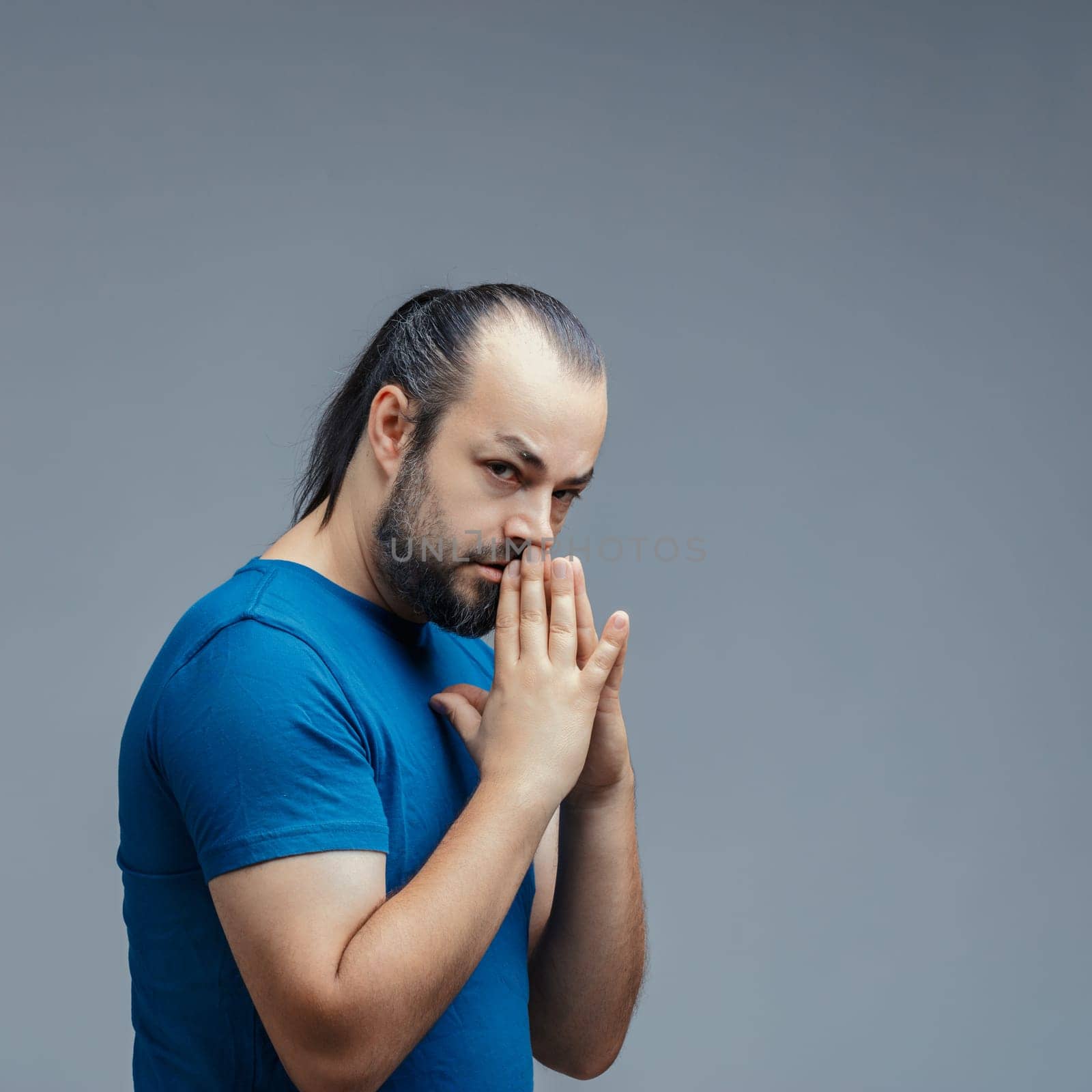 Pensive adult man with a beard and ponytail, wearing a blue t-shirt and holding his hands near his mouth, gazes seriously at the camera against a plain background