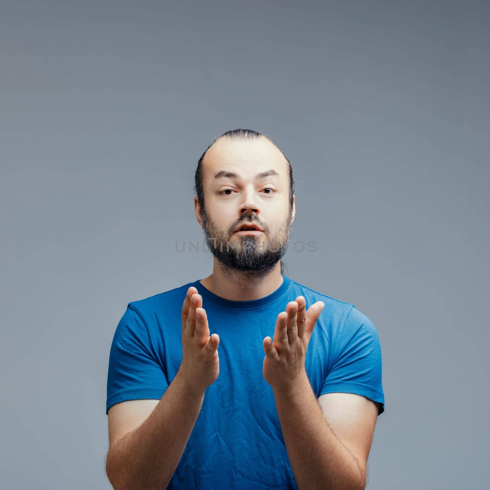 Caucasian man with beard and ponytail, wearing blue t-shirt, looking surprised while making welcome and agreement gestures with hands, isolated on gray background
