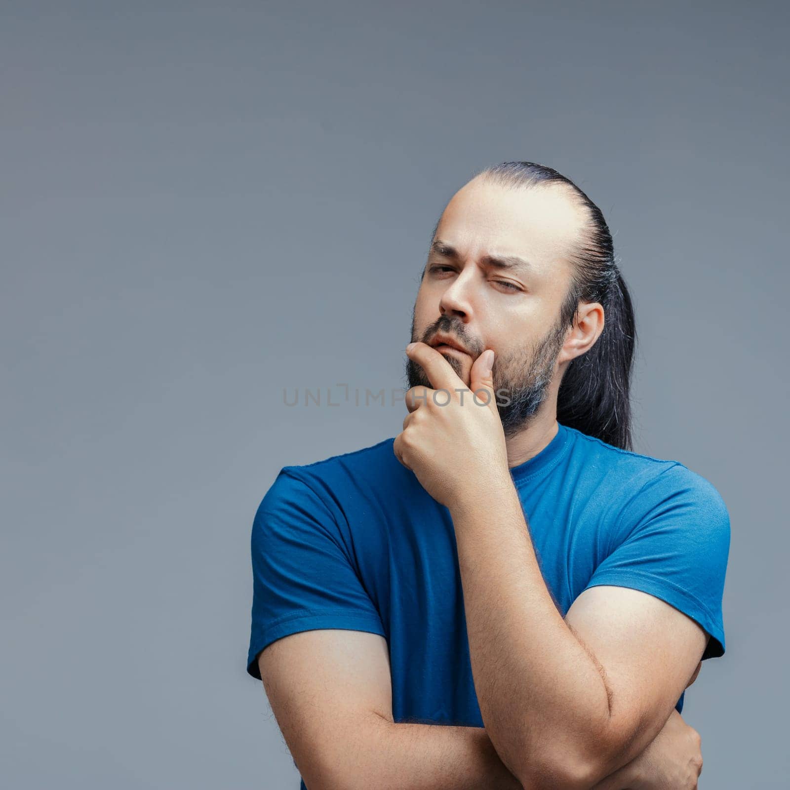 Studio shot of a thoughtful man with a suspicious expression, touching his chin with his hand, wearing a blue t-shirt on a gray background, conveying emotions, feelings, thoughts, and expressions