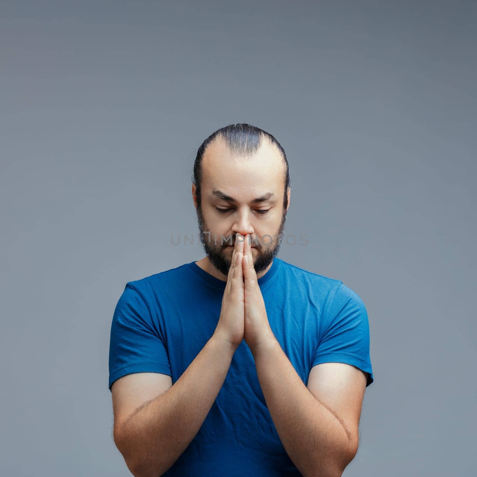 Contemplative man with clasped hands reflects in a moment of introspection, his serene expression conveying a sense of peace and mindfulness against a neutral gray backdrop