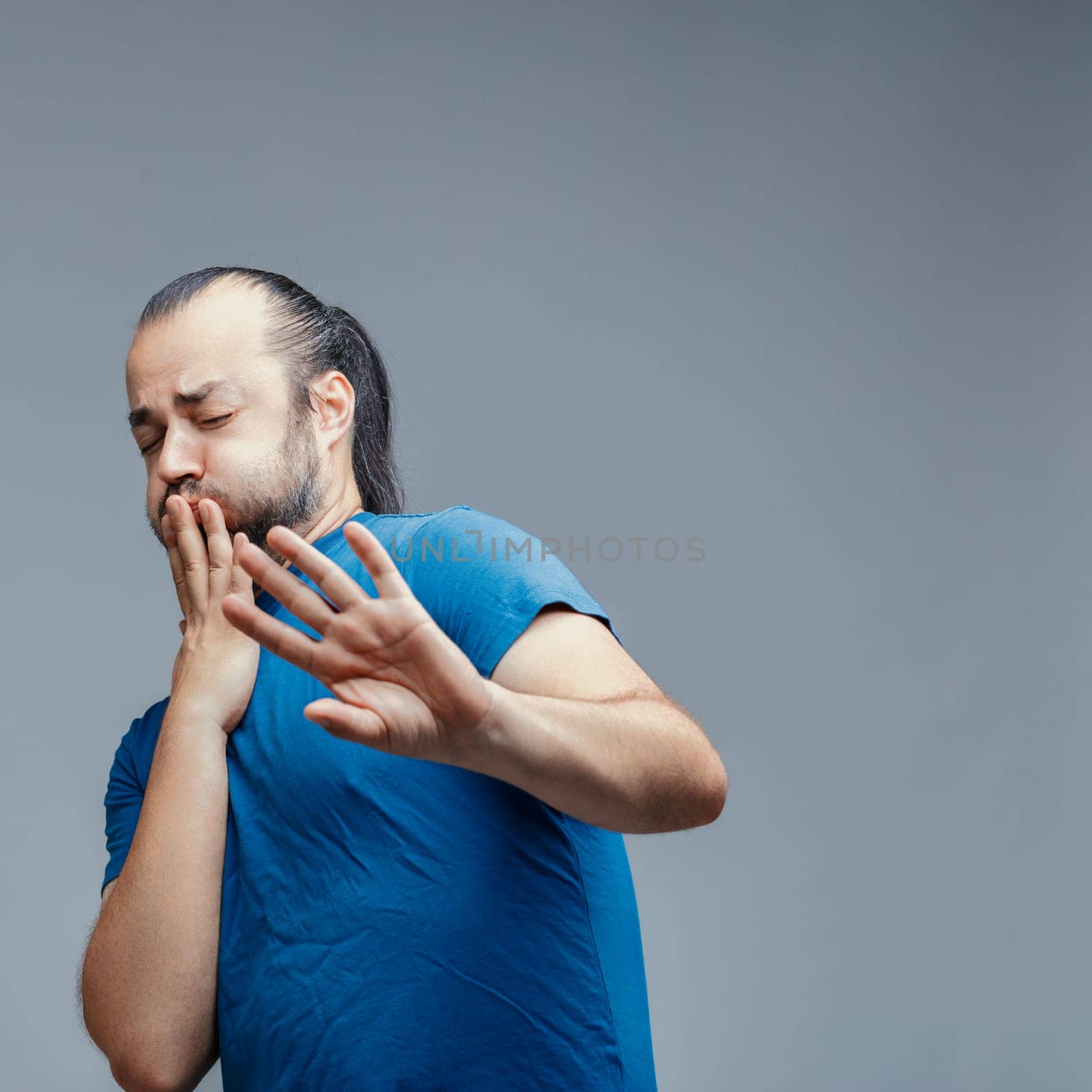 Young adult man with beard and long hair tied in a ponytail wearing a blue t-shirt is expressing disgust and covering his mouth with his hand while also pushing away with the other hand