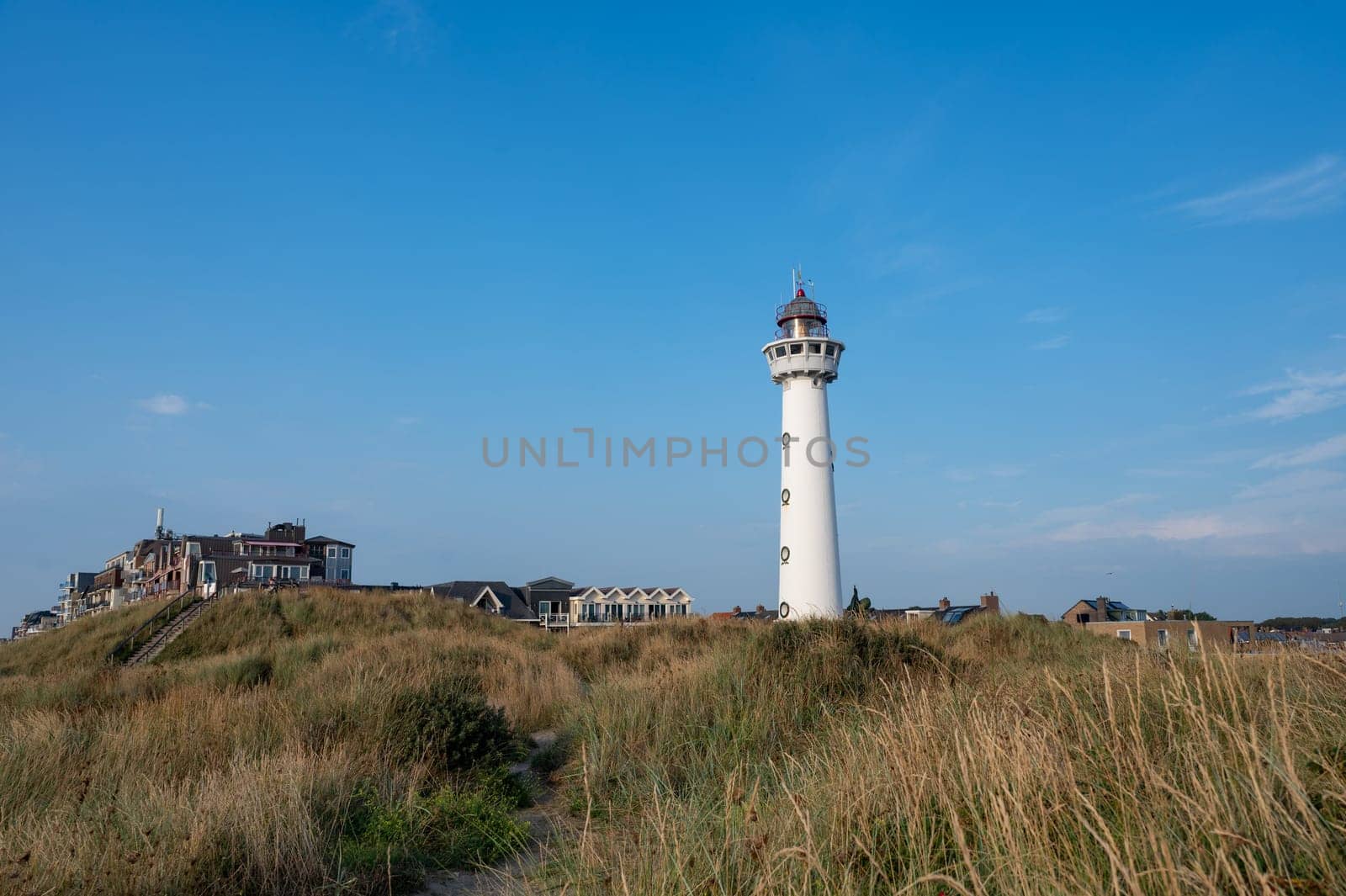 The approx. 28 m high white lighthouse J.C.J. van Speijk in Egmond aan Zee, North Holland, Netherlands with some of the town's houses with dune grass