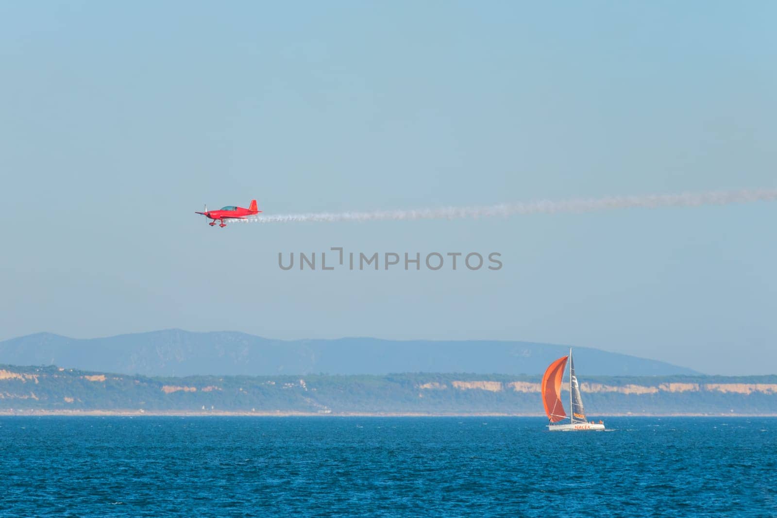 Red stunt aerobatic plane performing stunts in blue sky flying over sea with yacht boat. Concept of speed, difficulty, precision, risk