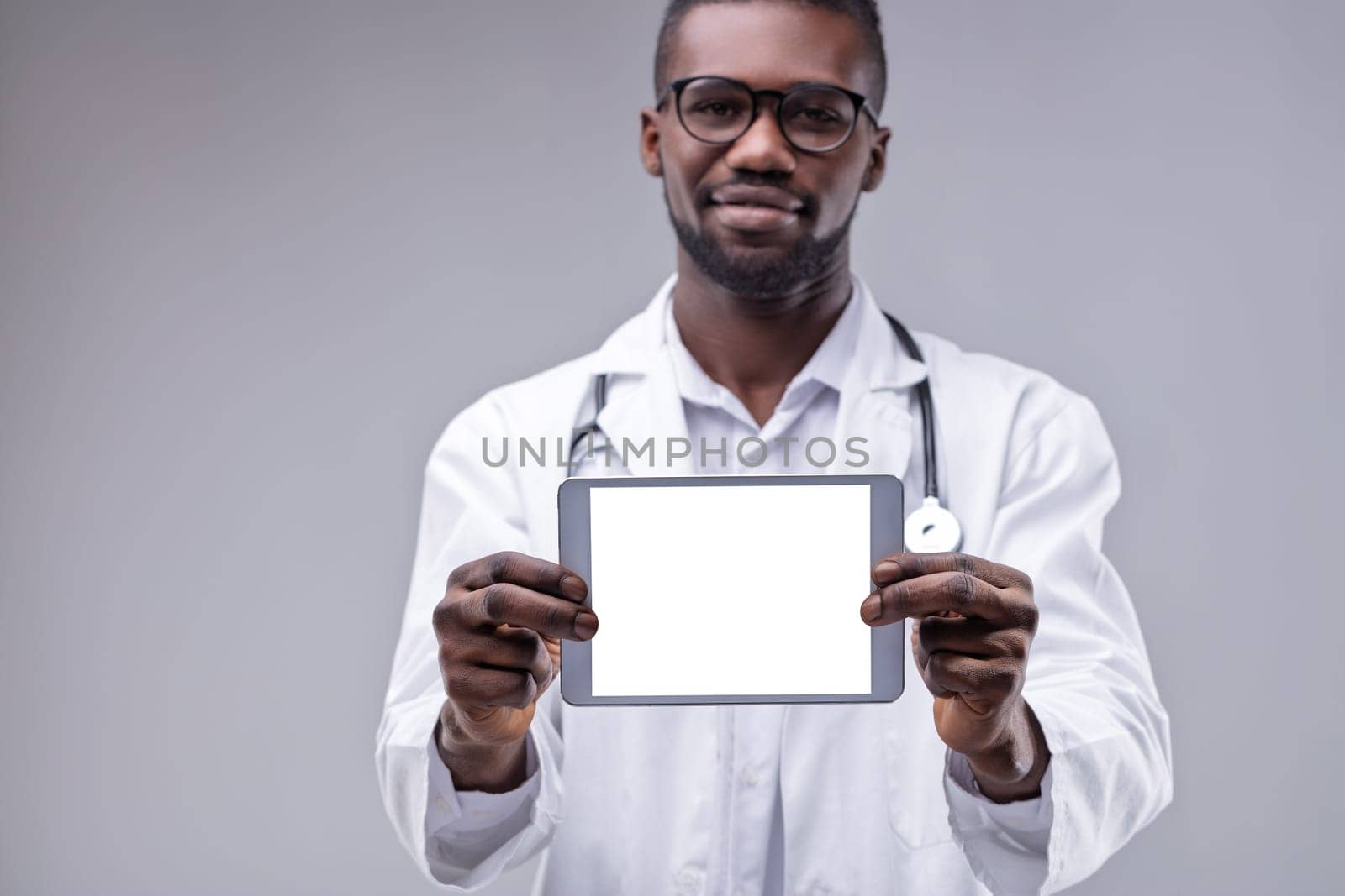 Smiling african american doctor wearing lab coat and stethoscope is holding a digital tablet with blank white screen, providing space for custom text or image