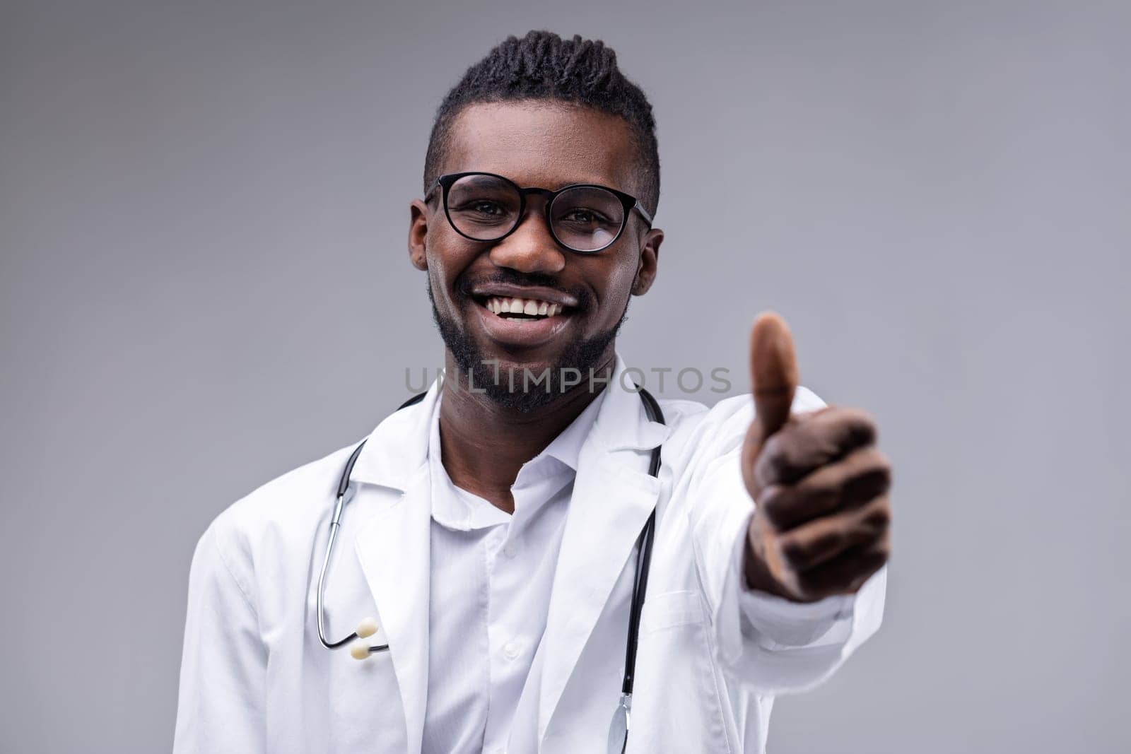 Young adult doctor, wearing a lab coat and stethoscope, smiling broadly while giving a thumbs up gesture, exuding confidence and positivity against a gray background