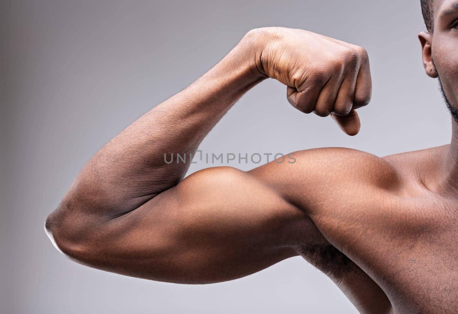 Close-up of a muscular black man flexing his impressive biceps, highlighting strength and fitness against a smooth gray backdrop, embodying power and determination in his physique