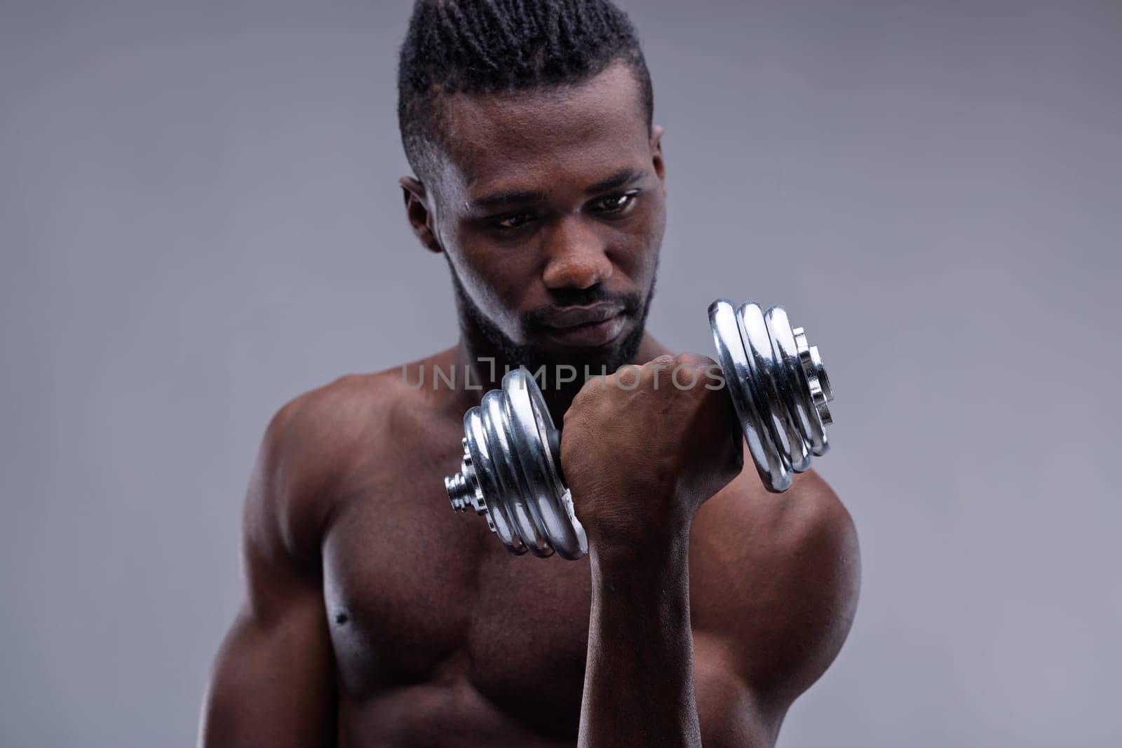 Muscular black man lifts a dumbbell with a determined expression, showcasing his strength and dedication to fitness against a gray studio backdrop