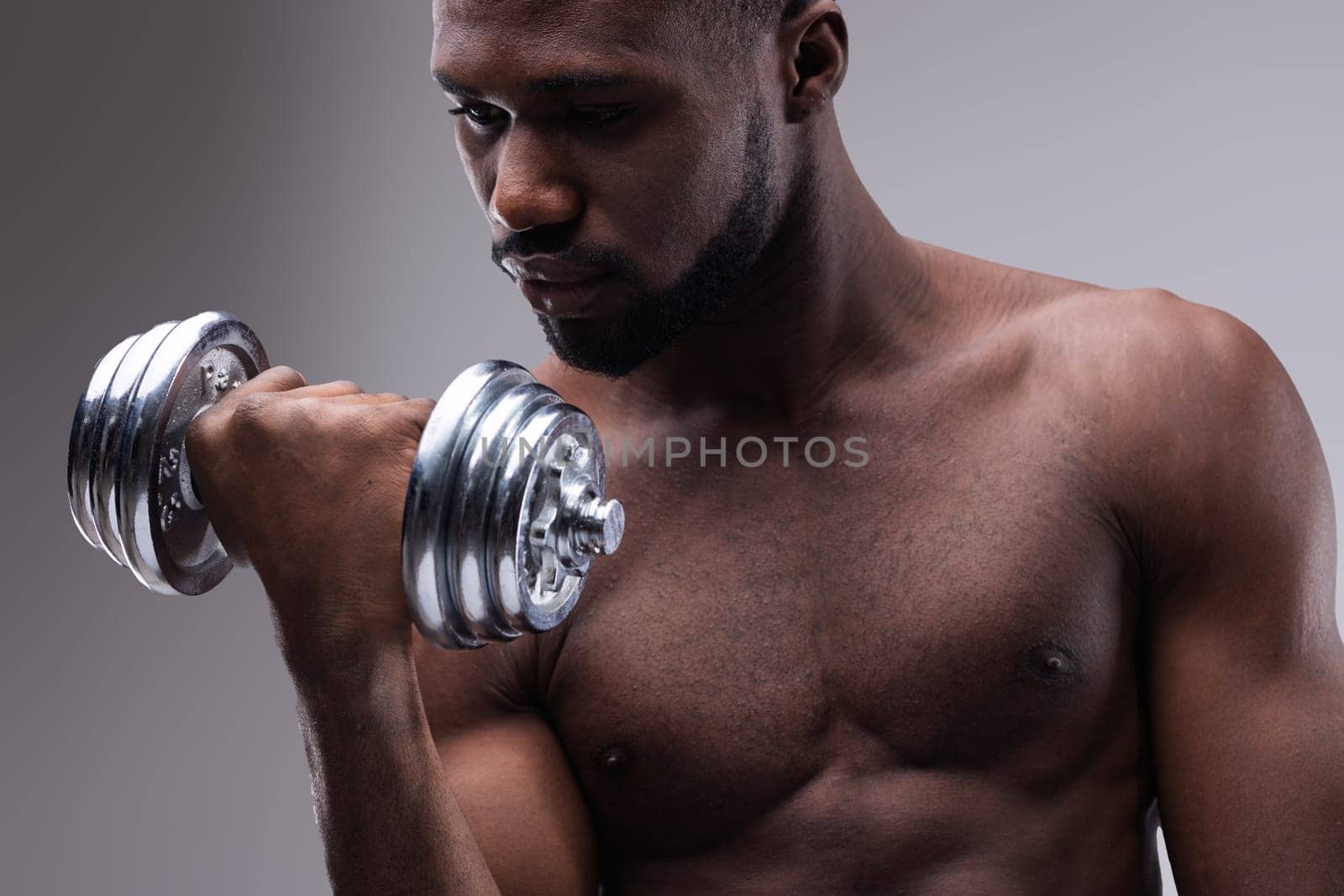 Strong young african american man lifting a dumbbell while intensely focusing on his arm, showcasing impressive muscles and strength during an energizing workout session