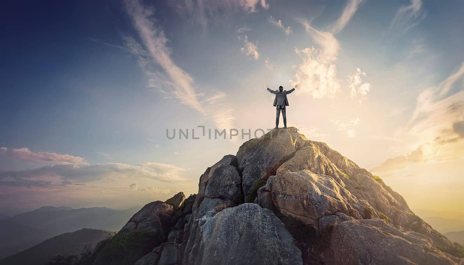 Businessman in suit standing triumphantly on the top of a mountain peak with arms raised up celebrating victory success. Achievement and reaching new heights concept