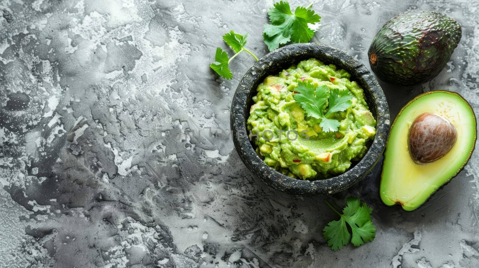 A top-down view of guacamole in a stone mortar, flanked by halved avocados and fresh cilantro. The composition highlights the ingredients' natural textures and colors