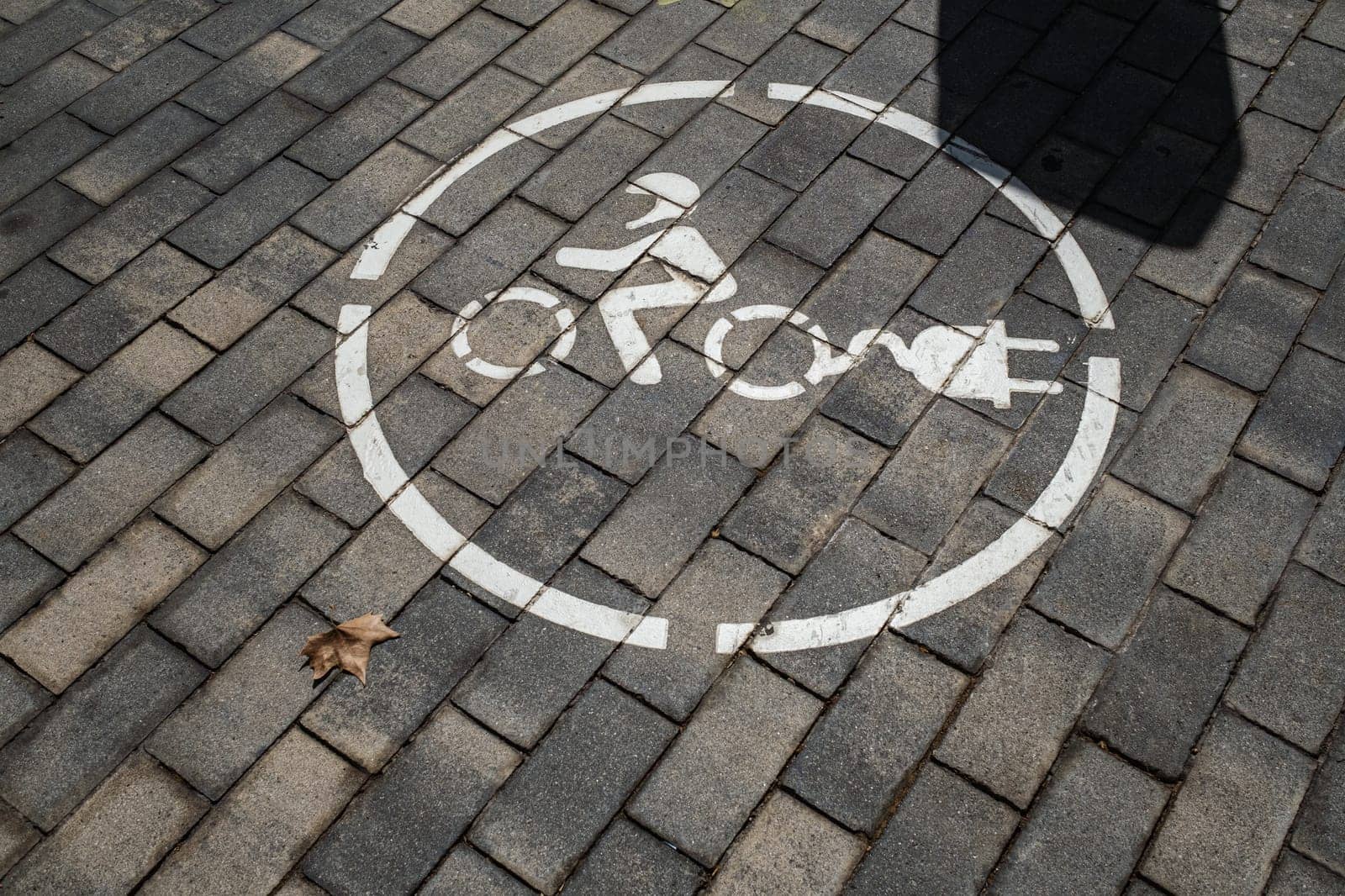 Service area for electric charging for motorcycles. Stylized drawing of a motorcyclist on two wheels with electric charging plug, circular outline, painted white. Background of concrete brick pavement.