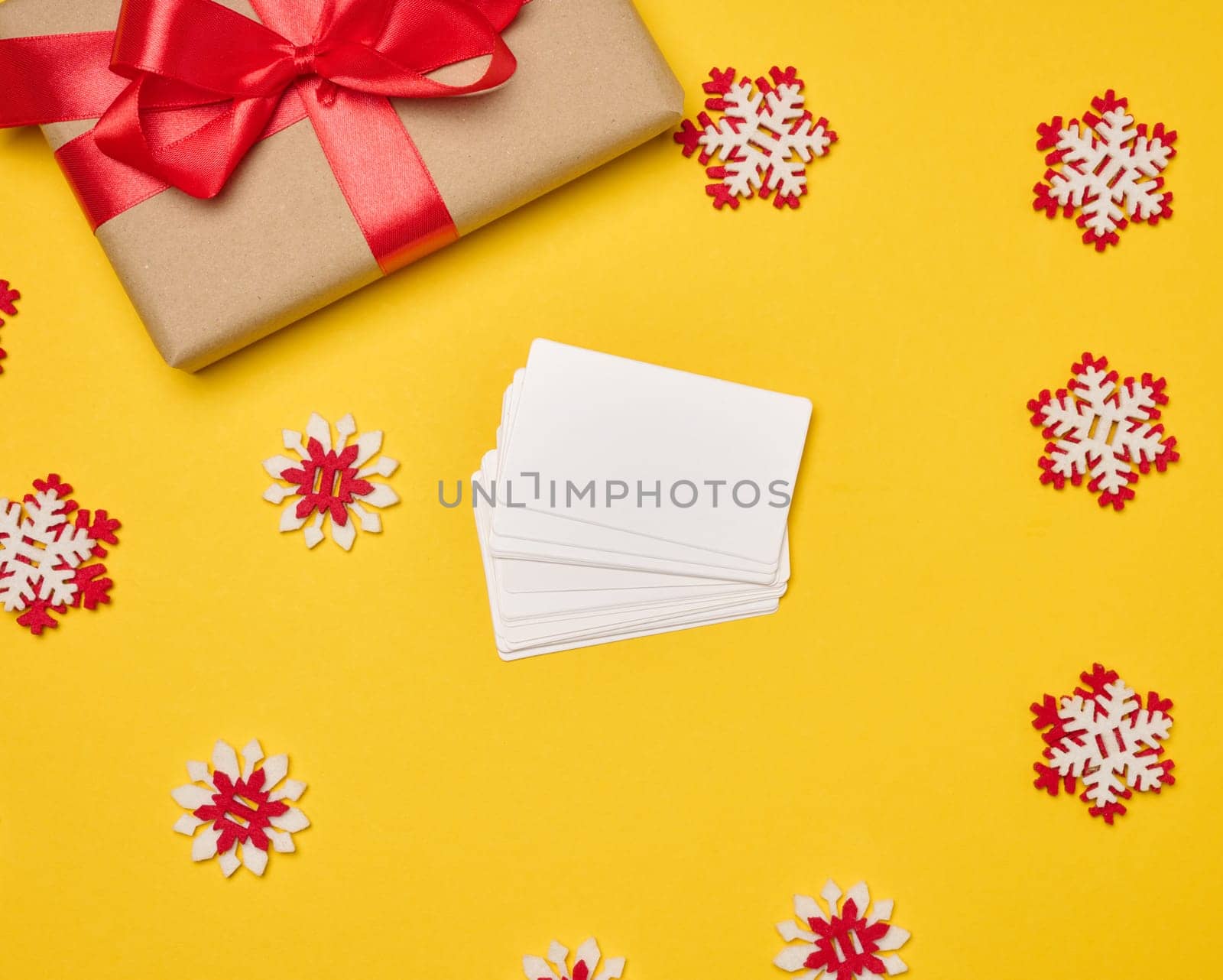 Christmas gift wrapped in red ribbon on yellow background, stack of white paper business cards. Top view